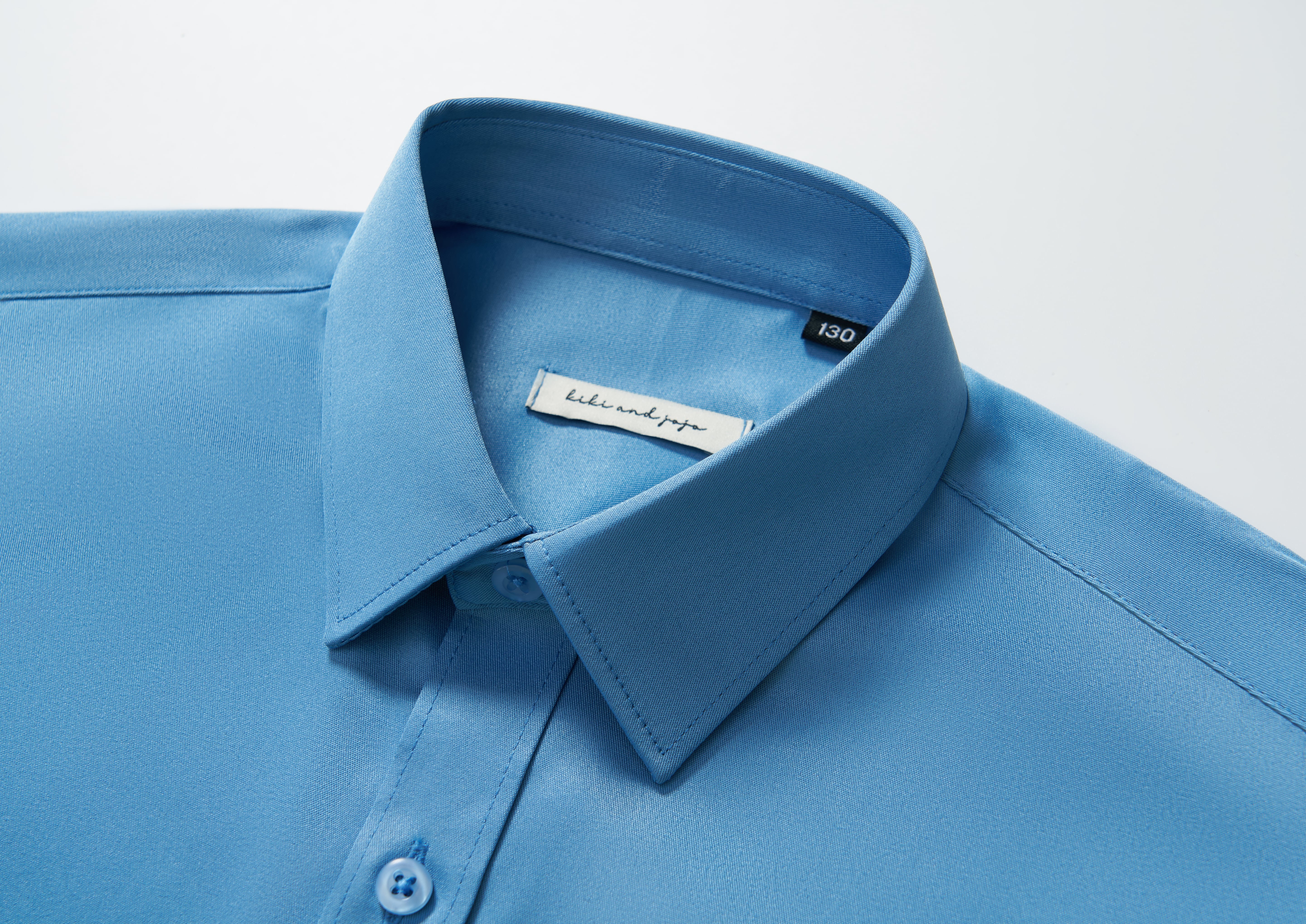 Close-up of the KIKI and JOJO Boys Sky Blue Dress Shirt, showing buttons and the brand label inside the collar, set against a white background.