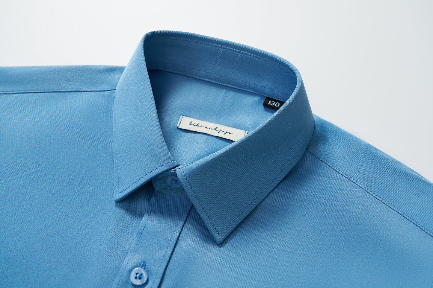 Close-up of the KIKI and JOJO Boys Sky Blue Dress Shirt, showing buttons and the brand label inside the collar, set against a white background.