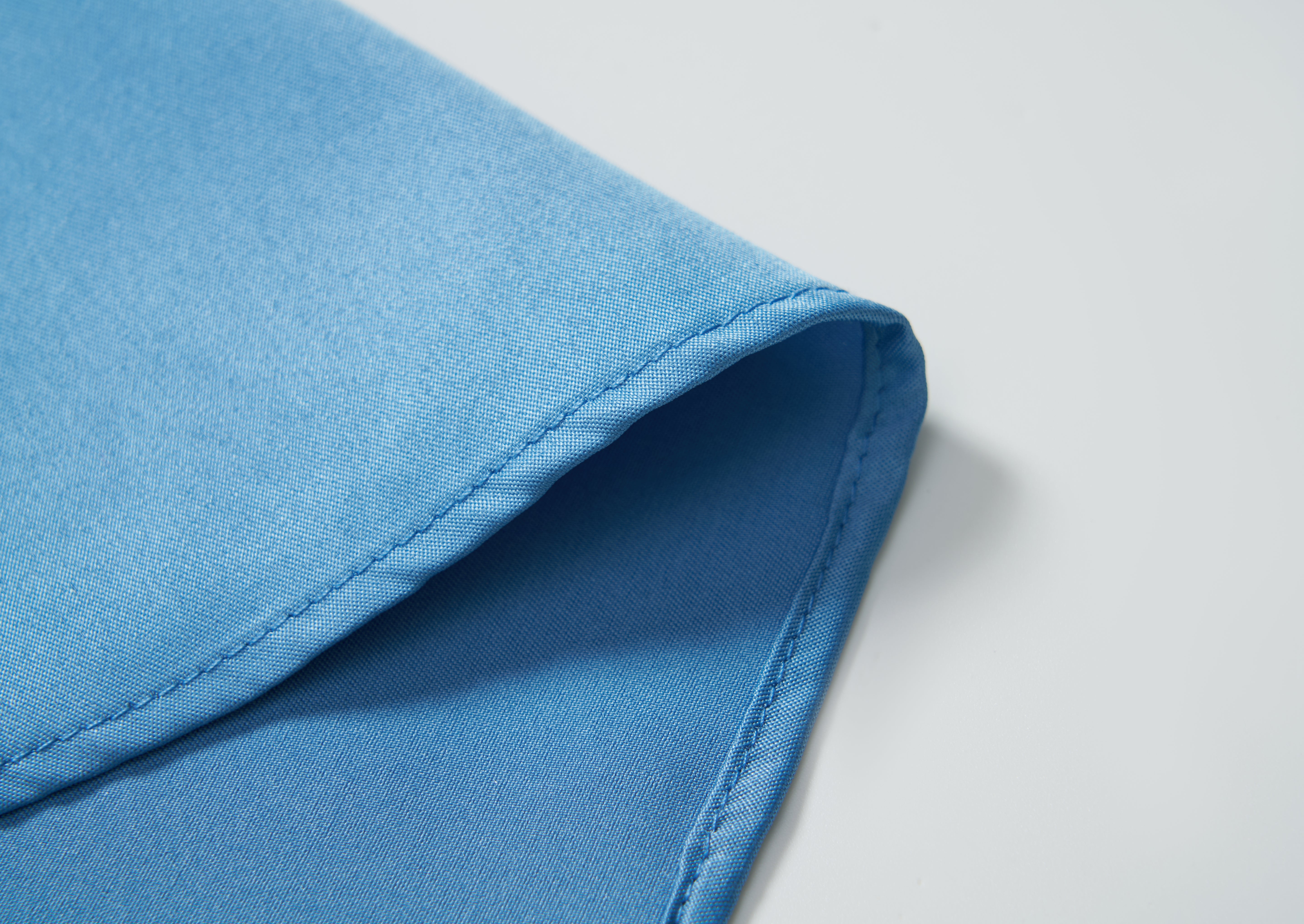 A close-up of the corner of the KIKI and JOJO Boys Sky Blue Dress Shirt highlights its crisp fabric, neatly folded on a white surface.