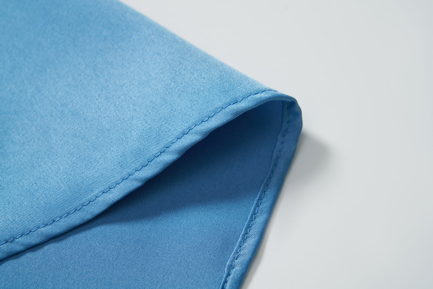 A close-up of the corner of the KIKI and JOJO Boys Sky Blue Dress Shirt highlights its crisp fabric, neatly folded on a white surface.