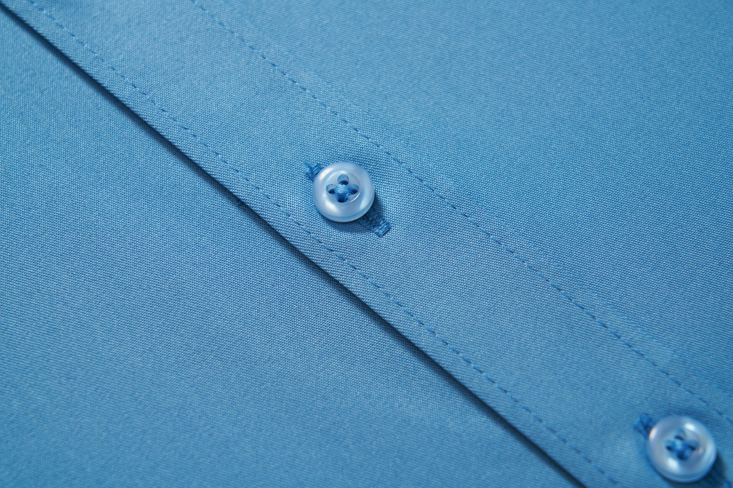 Close-up of KIKI and JOJO Boys Sky Blue Dress Shirt fabric, featuring visible stitching and two plastic buttons on the placket.