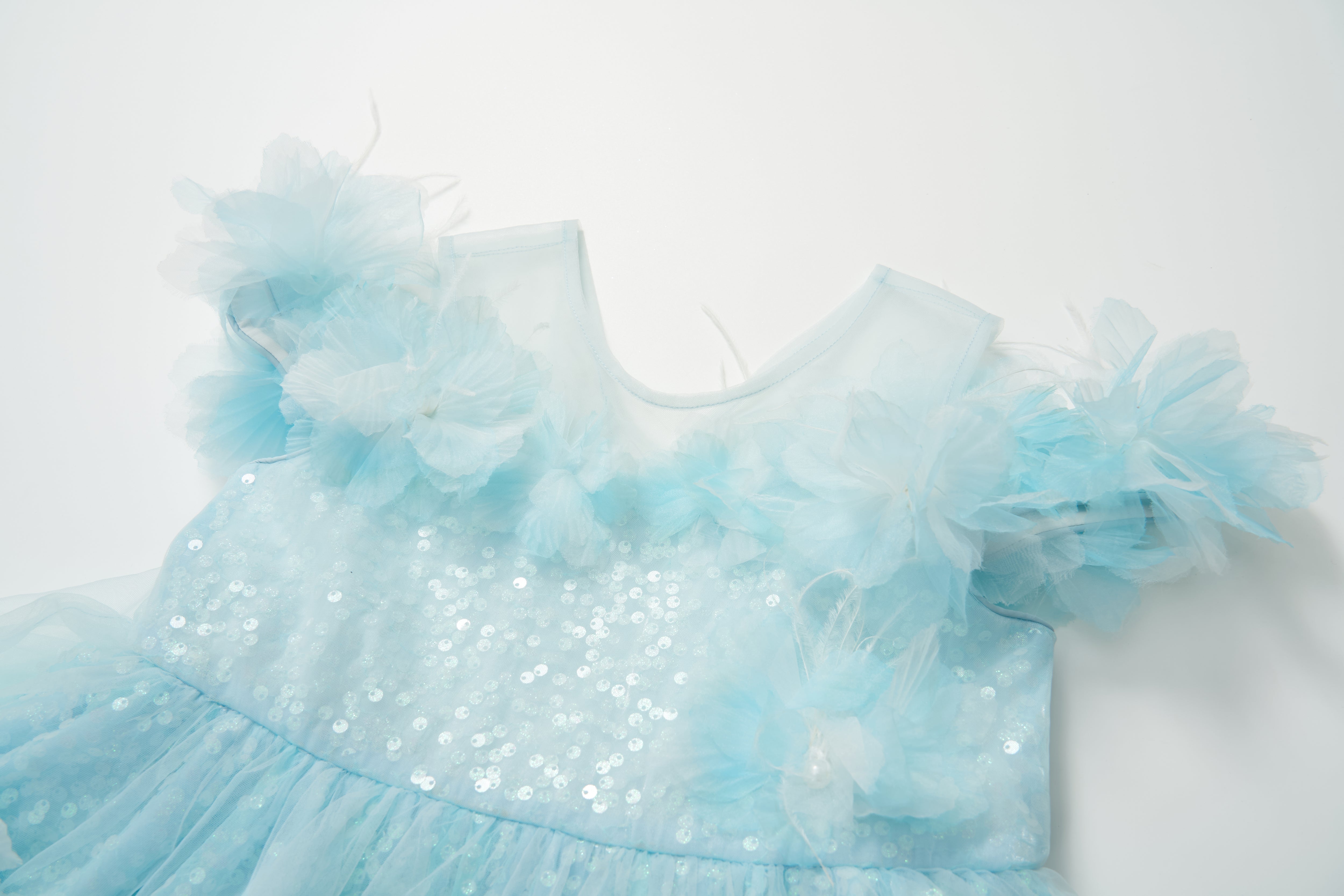 The Feathered Whisper Dress - Blue by KIKI and JOJO features light blue sequins, feather accents, and sheer floral shoulder details, displayed on a white background for an ethereal, elegant look.