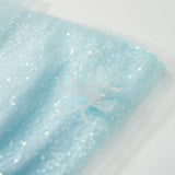 The Feathered Whisper Dress - Blue by KIKI and JOJO features light blue sequin fabric with a sheer overlay, accented by a fabric flower and feather embellishment, shown on a white background.