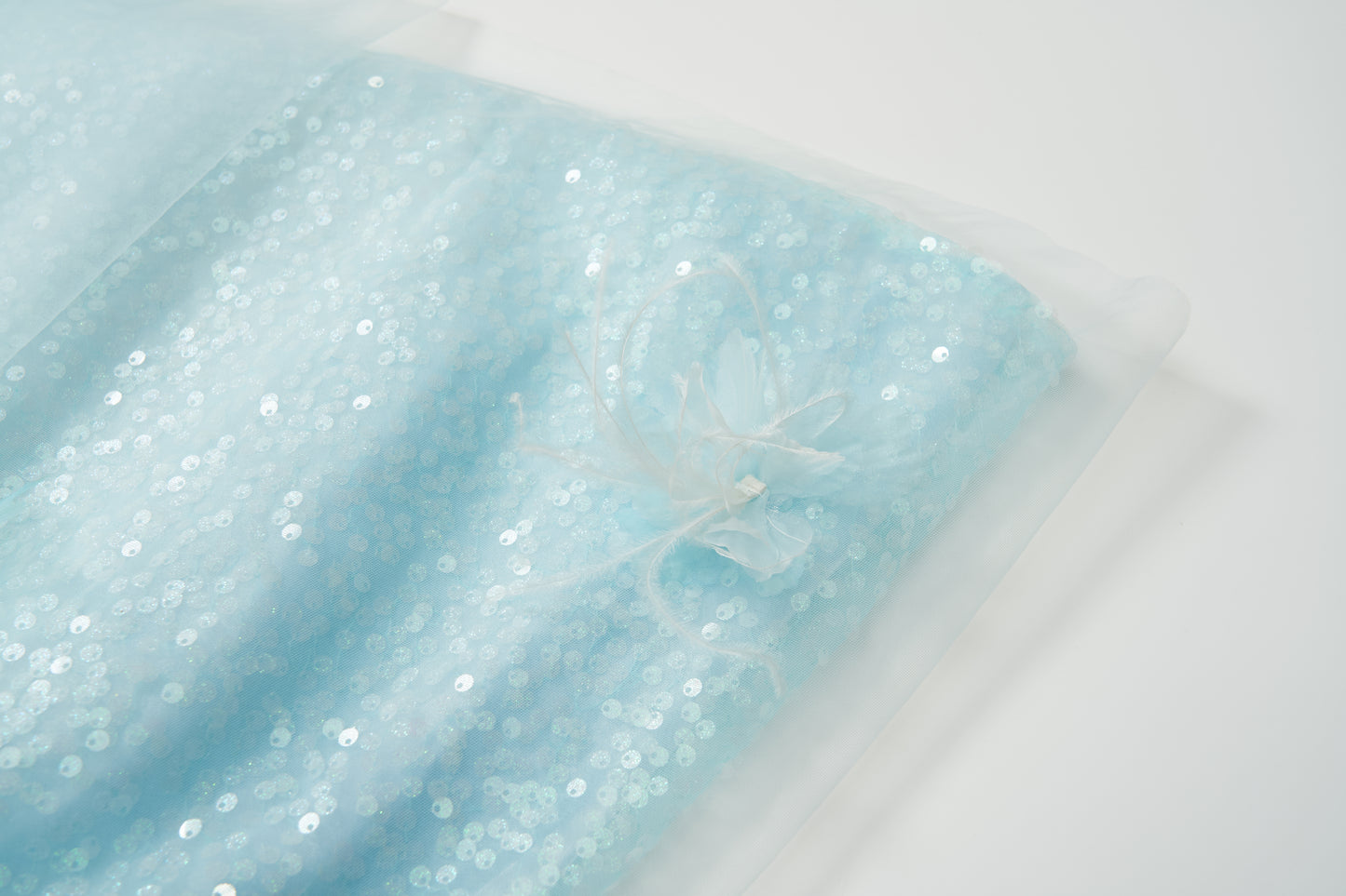 The Feathered Whisper Dress - Blue by KIKI and JOJO features light blue sequin fabric with a sheer overlay, accented by a fabric flower and feather embellishment, shown on a white background.