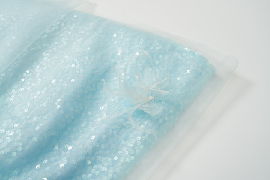 The Feathered Whisper Dress - Blue by KIKI and JOJO features light blue sequin fabric with a sheer overlay, accented by a fabric flower and feather embellishment, shown on a white background.