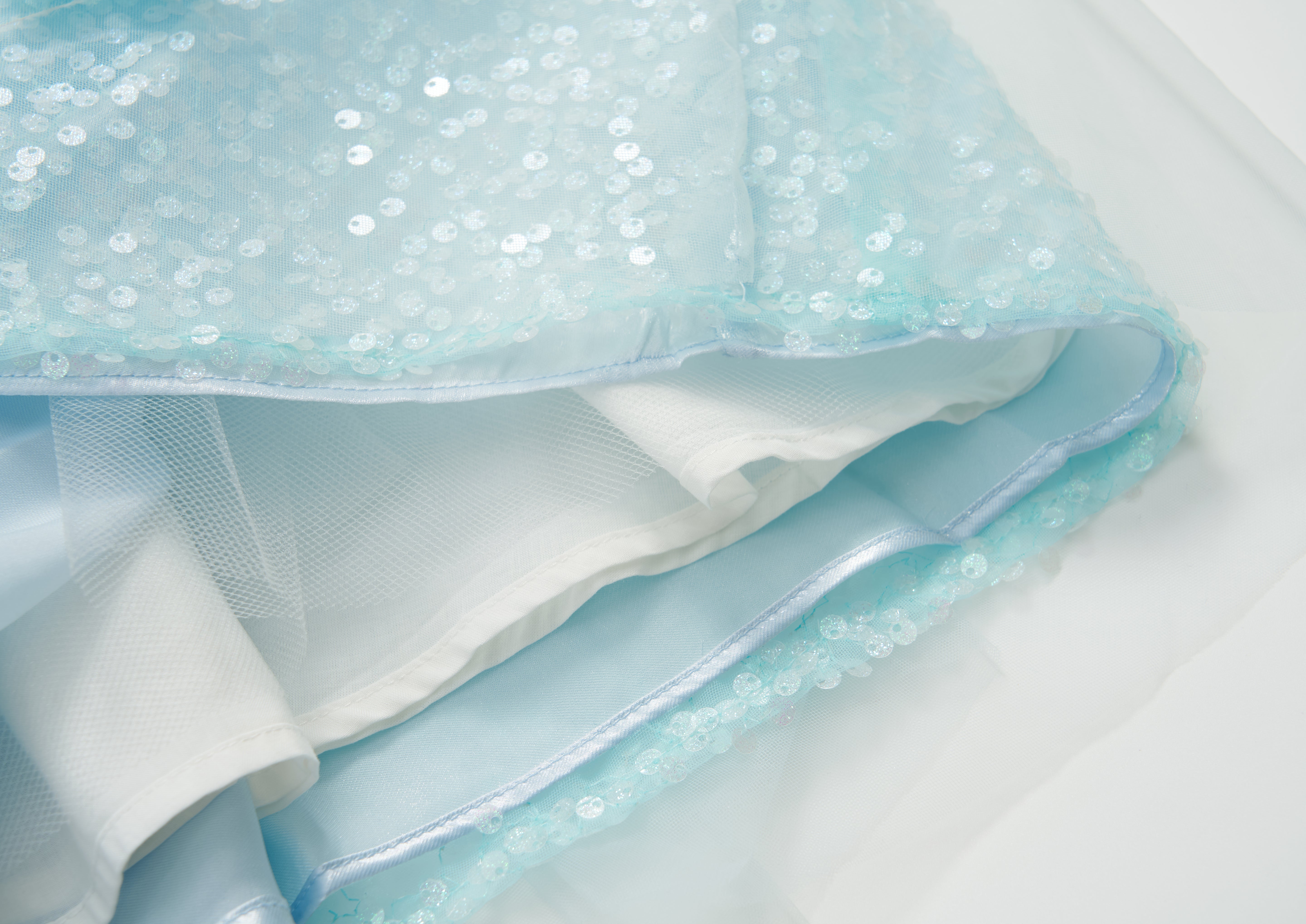 Close-up of the Feathered Whisper Dress - Blue by KIKI and JOJO: light blue sequined sheer fabric with white lining, radiating ethereal elegance—seen laid flat, highlighting its delicate off-shoulder design.