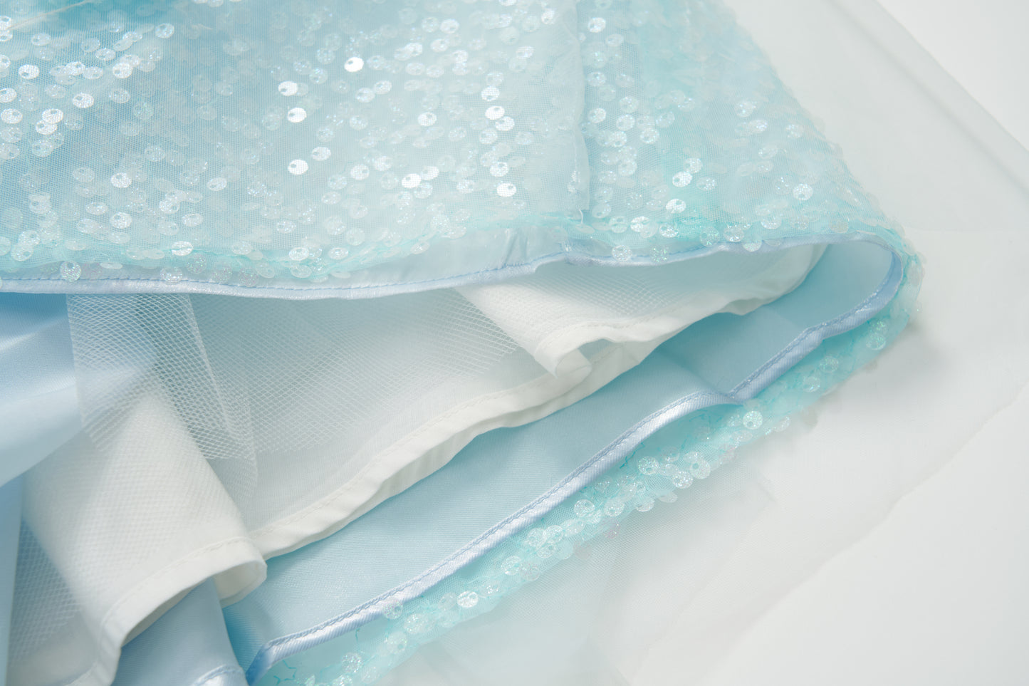 Close-up of the Feathered Whisper Dress - Blue by KIKI and JOJO: light blue sequined sheer fabric with white lining, radiating ethereal elegance—seen laid flat, highlighting its delicate off-shoulder design.