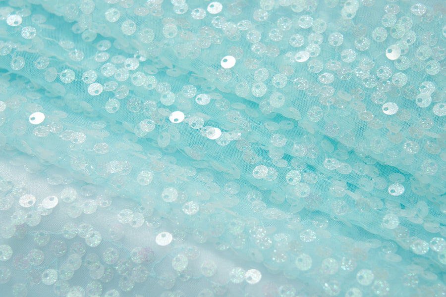 Close-up of the Feathered Whisper Dress - Blue by KIKI and JOJO, featuring pale blue sheer fabric adorned with round iridescent sequins for a shimmering, ethereal look—ideal for an off-shoulder statement.