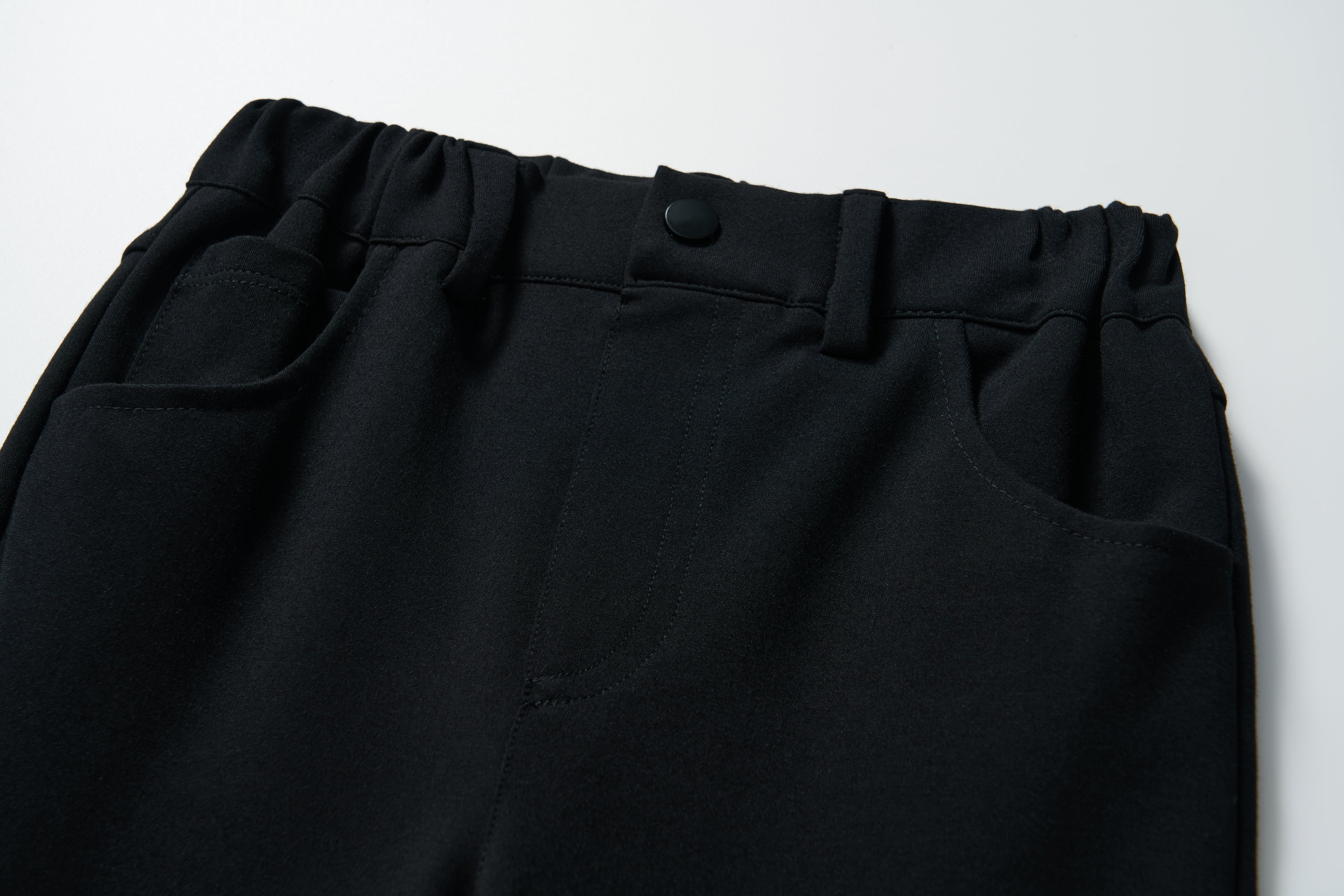 Close-up of the waistband and pockets of KIKI and JOJO's Petal Whisper Dress Pants, featuring flared black legs, stretchy fabric, an elastic waist, and button closure, shown on a white background.