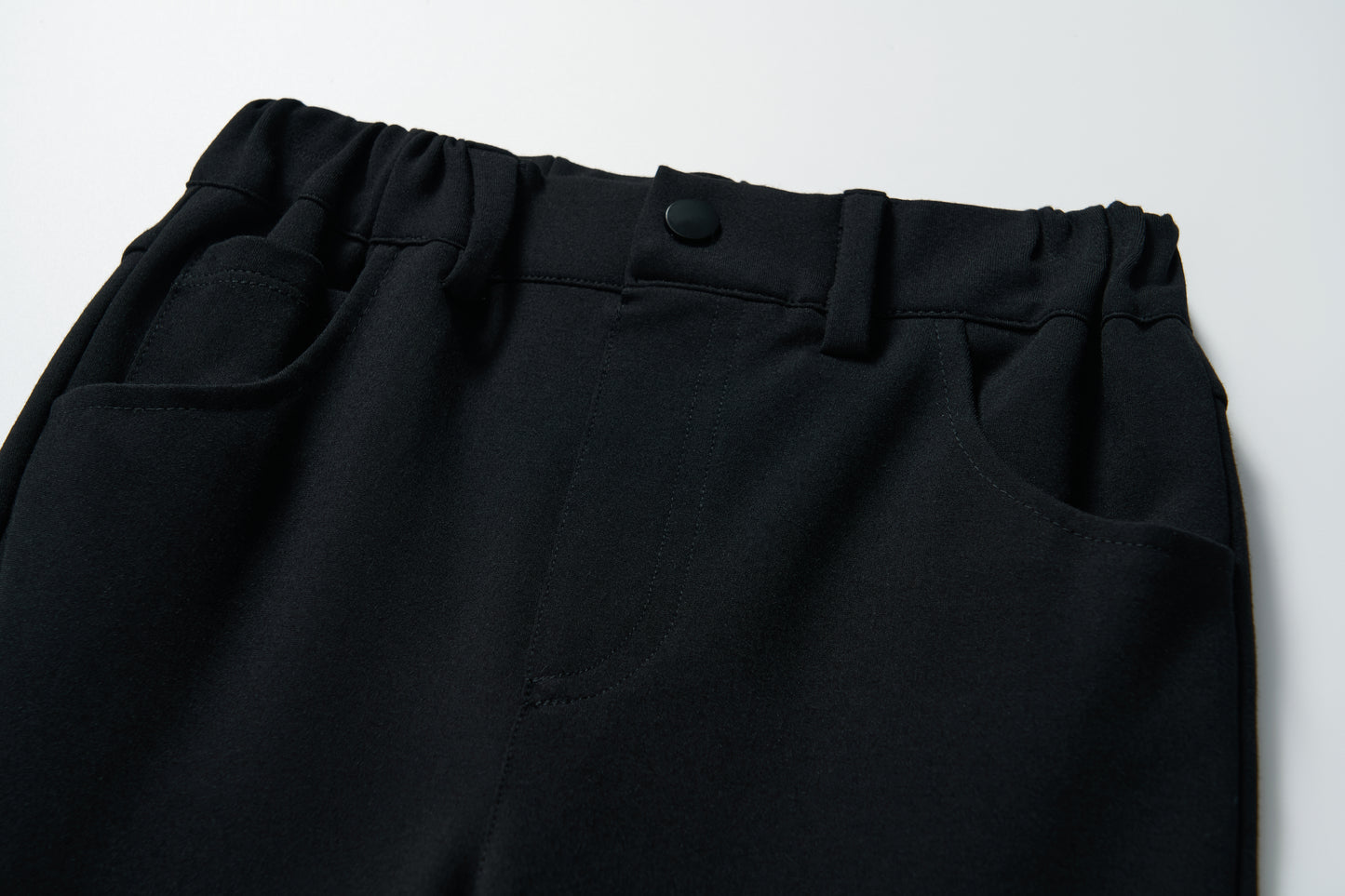 Close-up of the waistband and pockets of KIKI and JOJO's Petal Whisper Dress Pants, featuring flared black legs, stretchy fabric, an elastic waist, and button closure, shown on a white background.