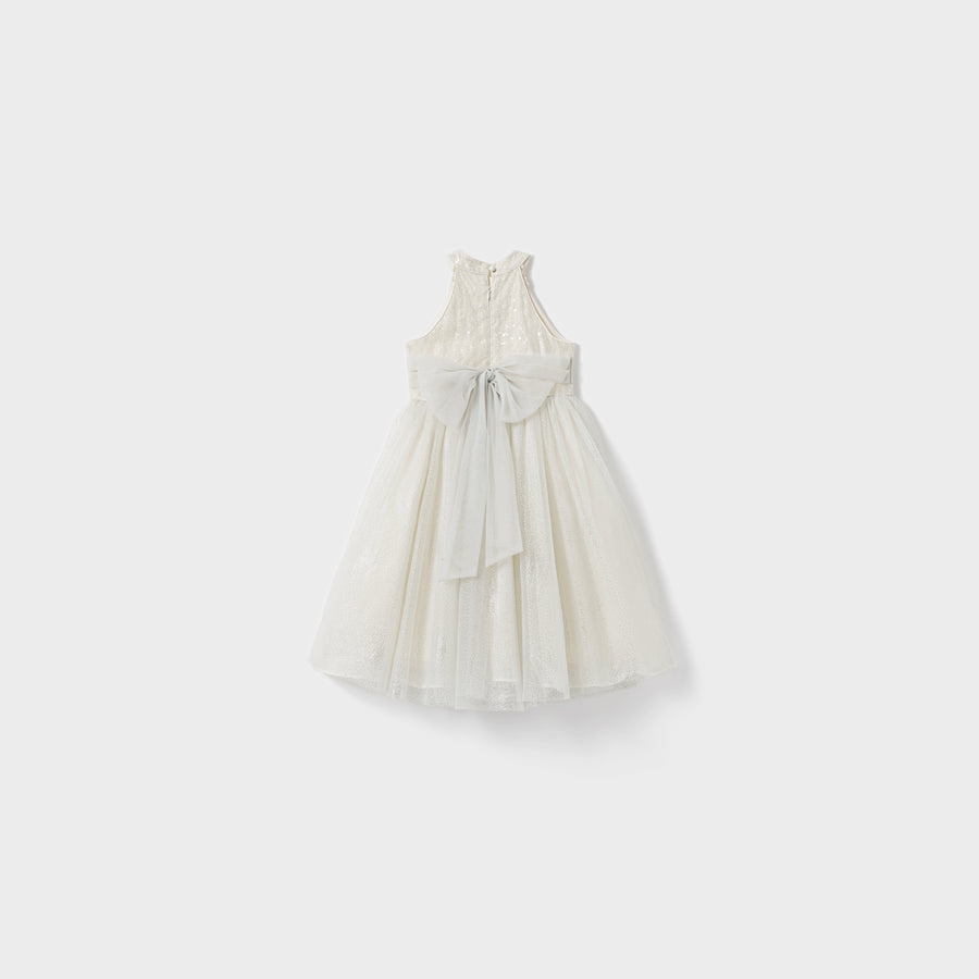 The Gilded Whisper Dress by KIKI and JOJO is a sleeveless white lace children's dress, featuring a large light gray back bow. Its classic beaded tassel-inspired design is beautifully displayed against a plain white background.
