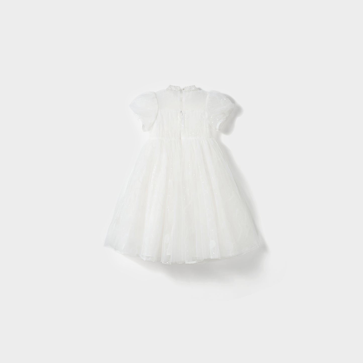 The Whisper Bloom Dress by KIKI and JOJO is a white, knee-length mini dress with short sleeves, floral embroidery, a tulle overlay, and subtle bead accents, shown against a plain light background.