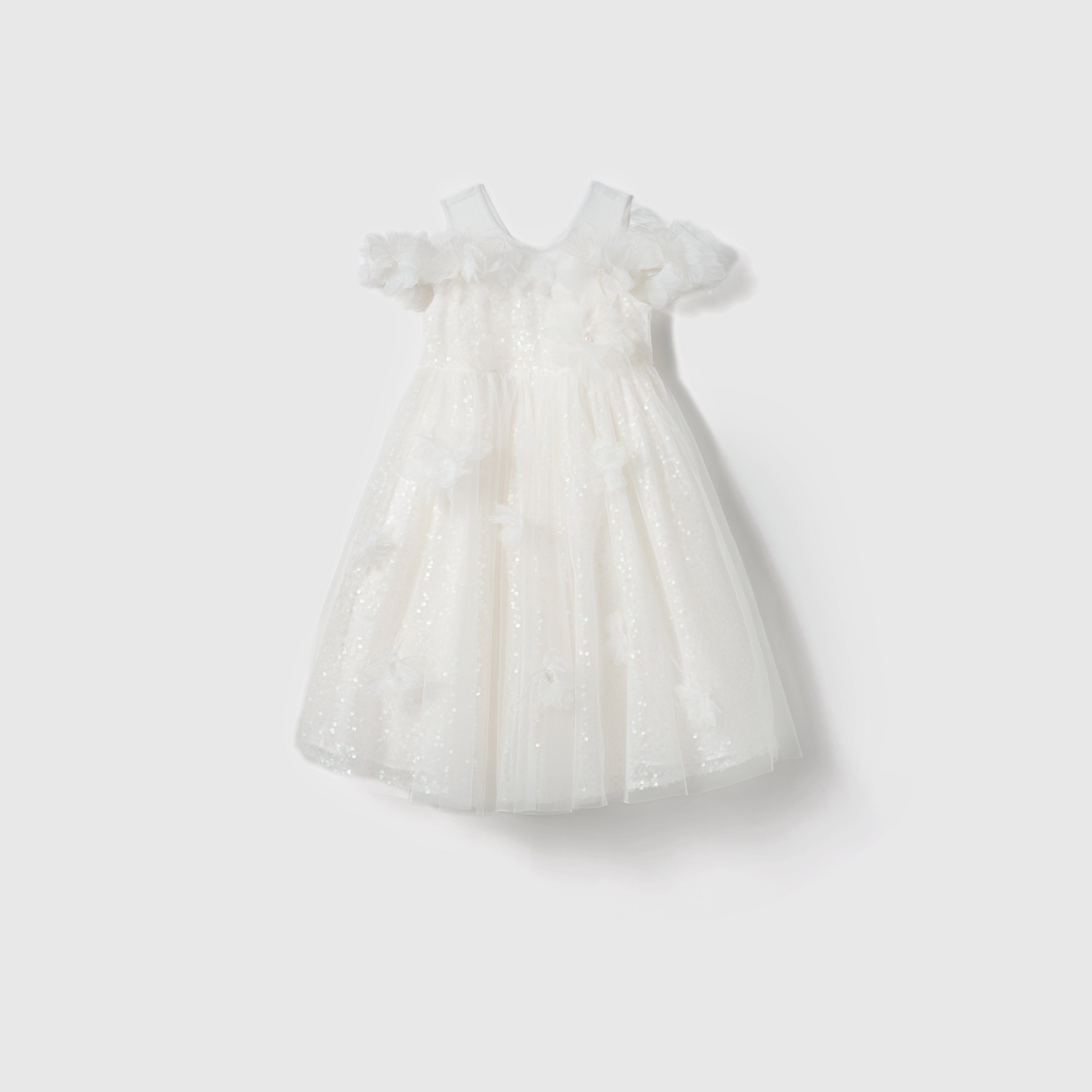 The Feathered Whisper Dress by KIKI and JOJO is a white sleeveless off-shoulder A-line dress for girls, featuring a tulle overlay, floral appliqués, and ruffle details, shown on a plain light background.