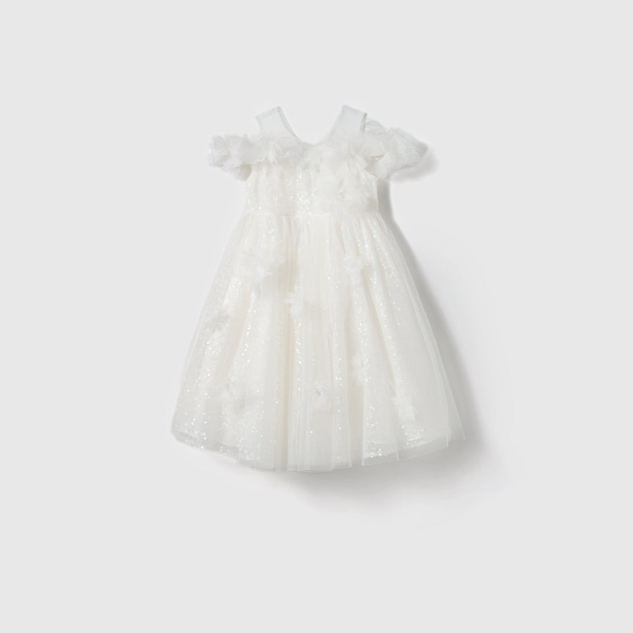 The Feathered Whisper Dress by KIKI and JOJO is a white sleeveless off-shoulder A-line dress for girls, featuring a tulle overlay, floral appliqués, and ruffle details, shown on a plain light background.