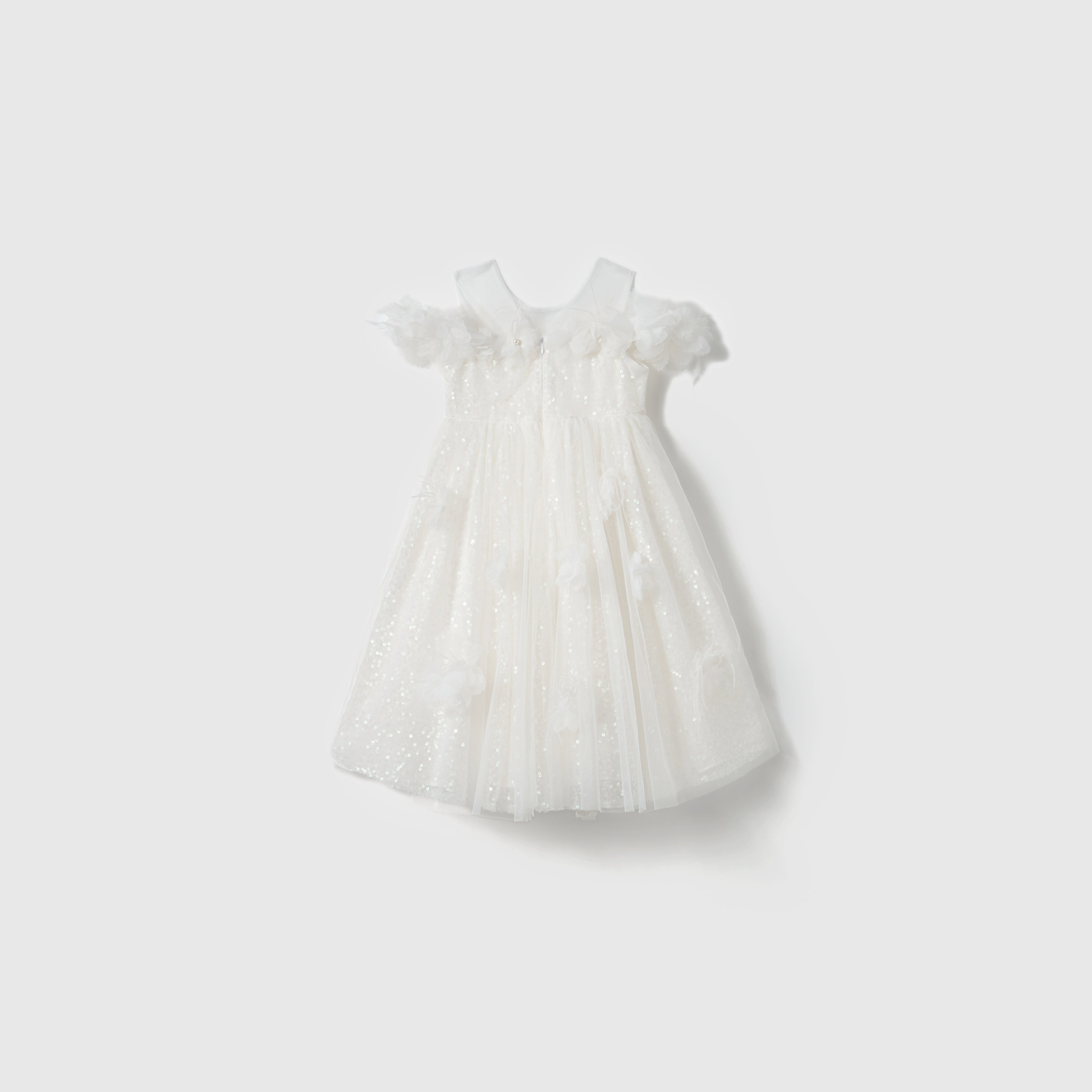 The Feathered Whisper Dress by KIKI and JOJO is a white, short-sleeved, knee-length dress with a tulle overlay and sequin floral appliqués. Subtle glitter details add elegance, shown on a simple light background.