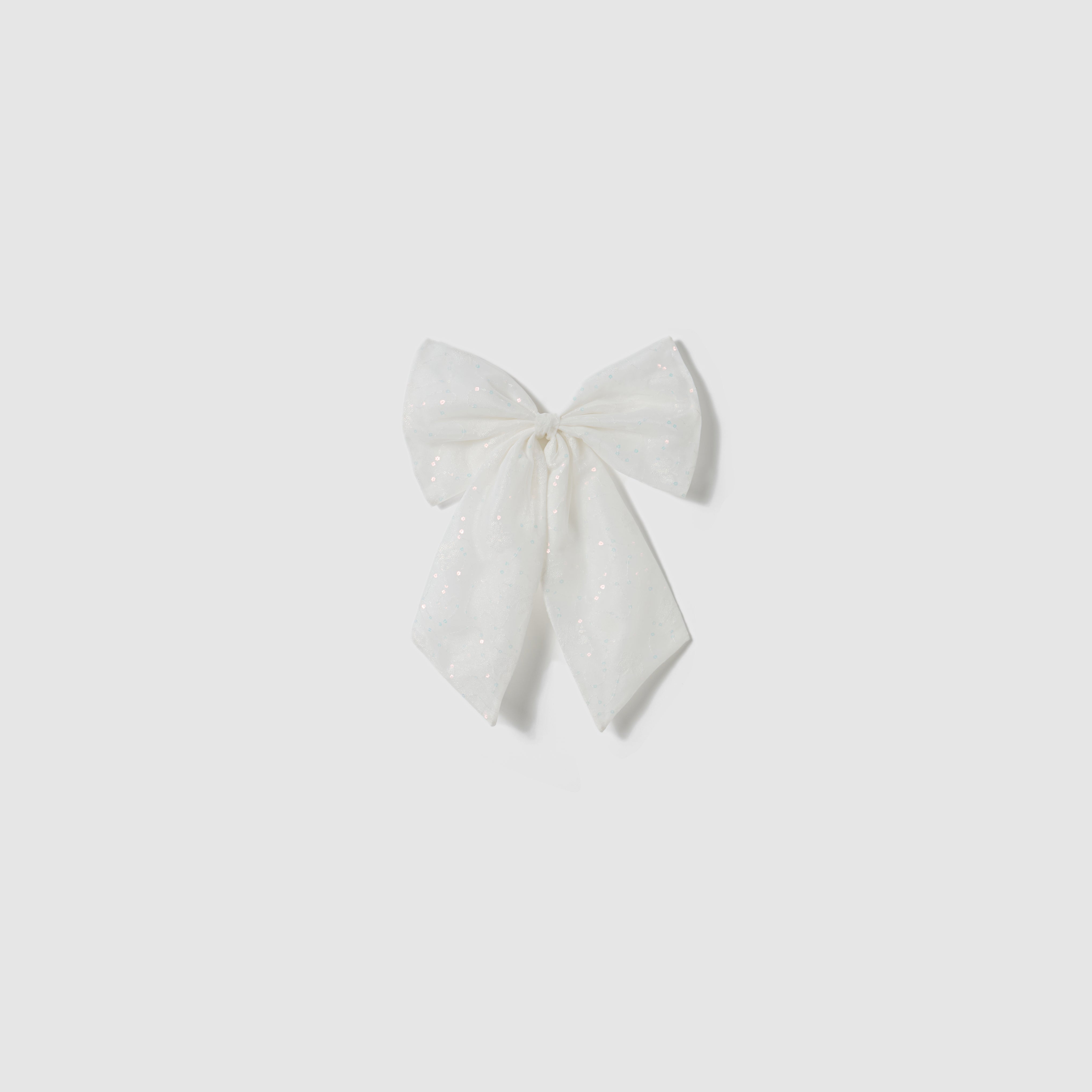 A large white fabric bow with subtle glitter details, inspired by the elegant Butterfly Whispers Dress from KIKI and JOJO, is displayed against a plain light gray background.