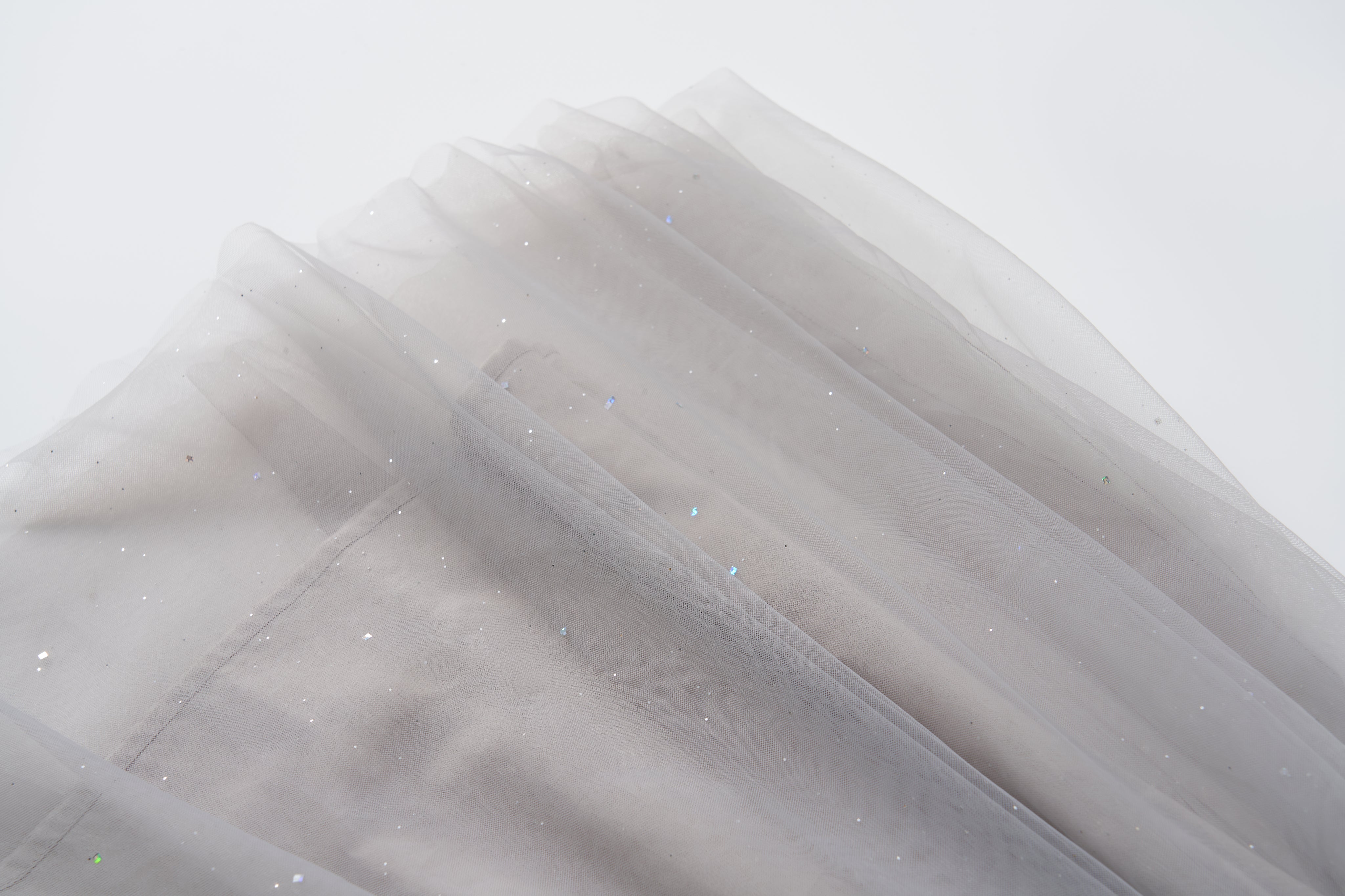 A close-up of soft, sheer light gray fabric with subtle glittery specks, draped in layers against a white background—ideal for creating the enchanting Dusk Whisper Dress by KIKI and JOJO.