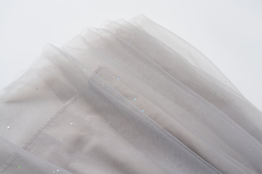 A close-up of soft, sheer light gray fabric with subtle glittery specks, draped in layers against a white background—ideal for creating the enchanting Dusk Whisper Dress by KIKI and JOJO.