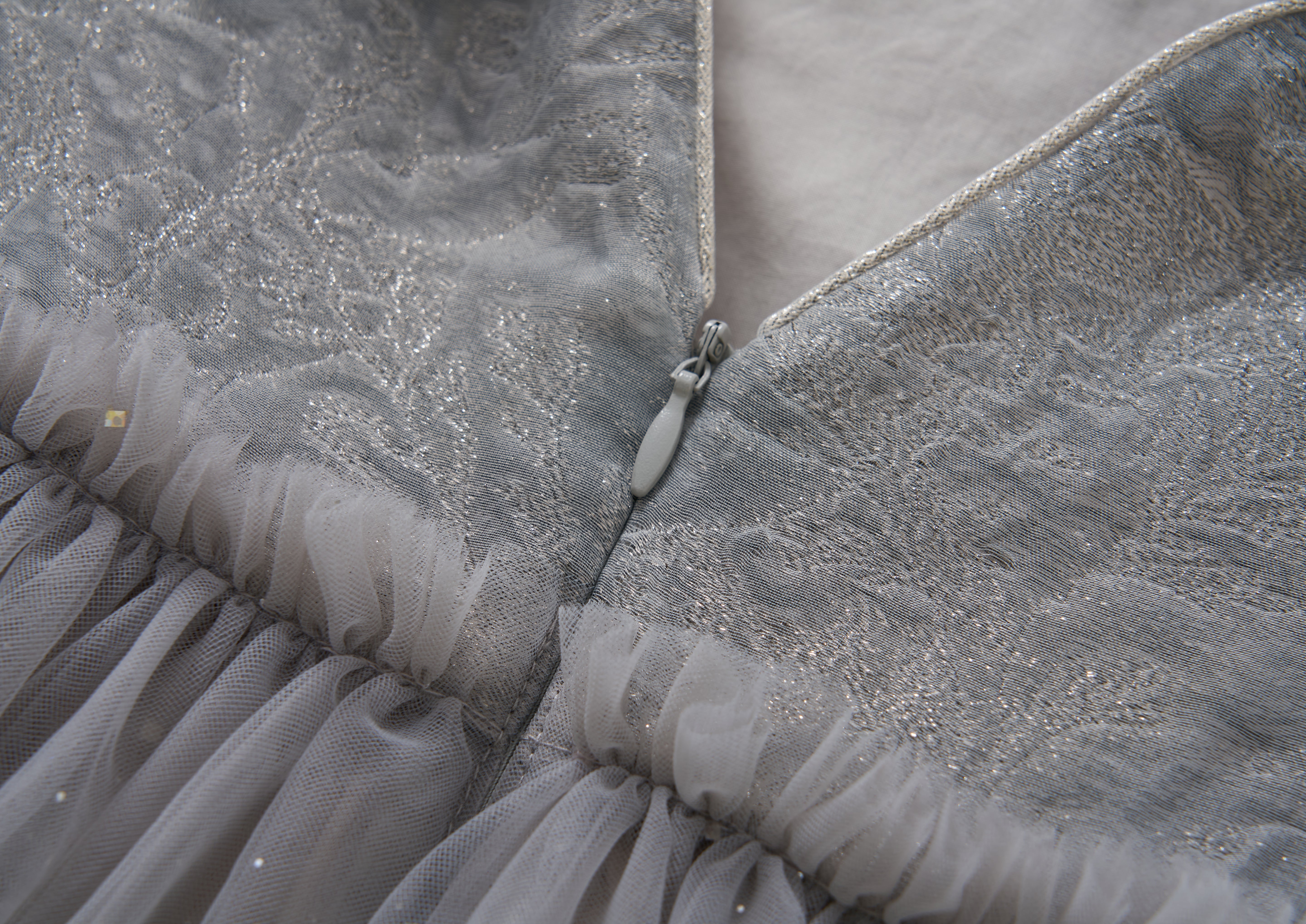 Close-up of the Dusk Whisper Dress by KIKI and JOJO, showcasing its glittering silver tulle, textured fabric, partially unzipped zipper revealing a soft lining, and gathered tulle detailing at the hem.