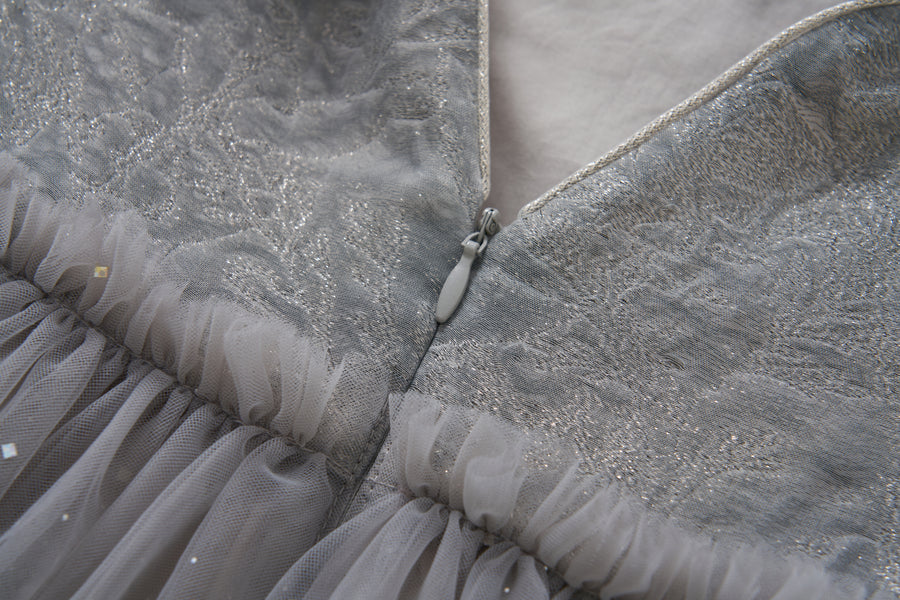 Close-up of the Dusk Whisper Dress by KIKI and JOJO, showcasing glittering silver tulle fabric, delicate tulle ruffles, and a partially unzipped silver zipper at the back.