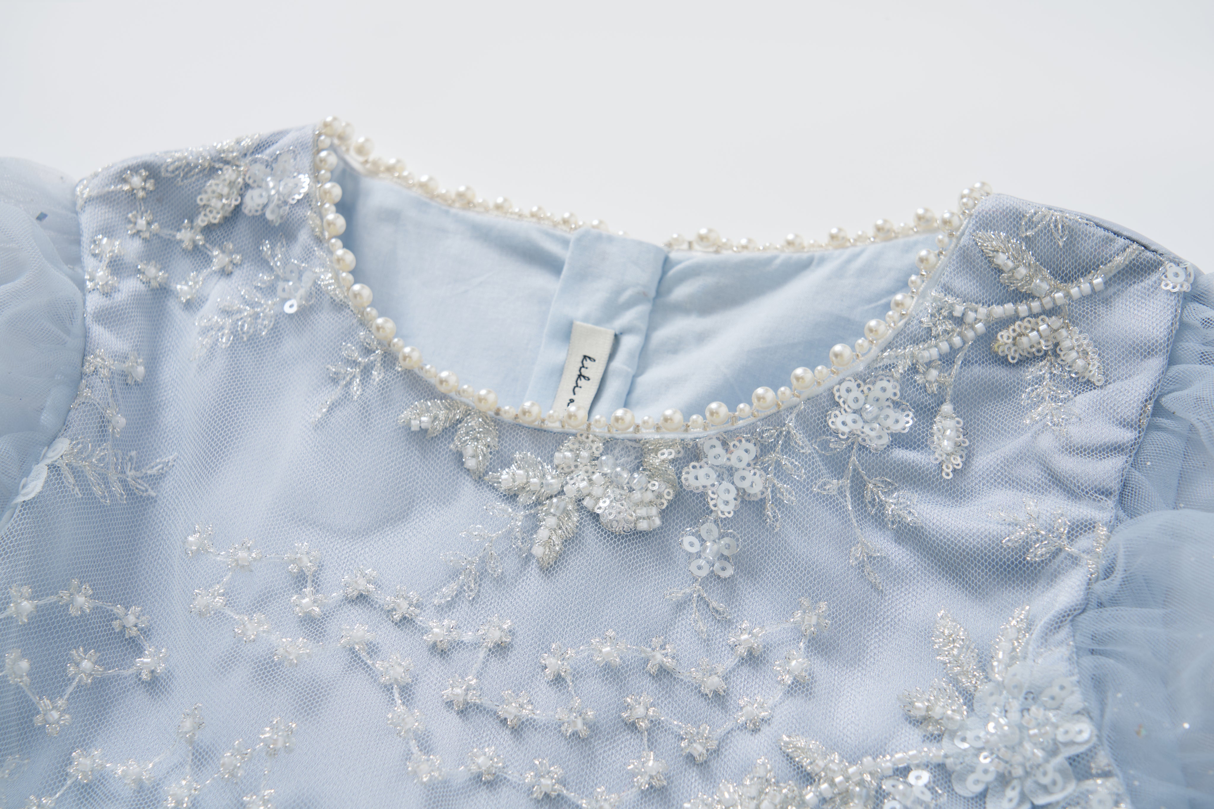 Close-up of the Mist Whisper Dress by KIKI and JOJO—an A-line light blue dress with hand-embroidered florals, sequins, sheer tulle sleeves, and a neckline finished with a delicate pearl trim.