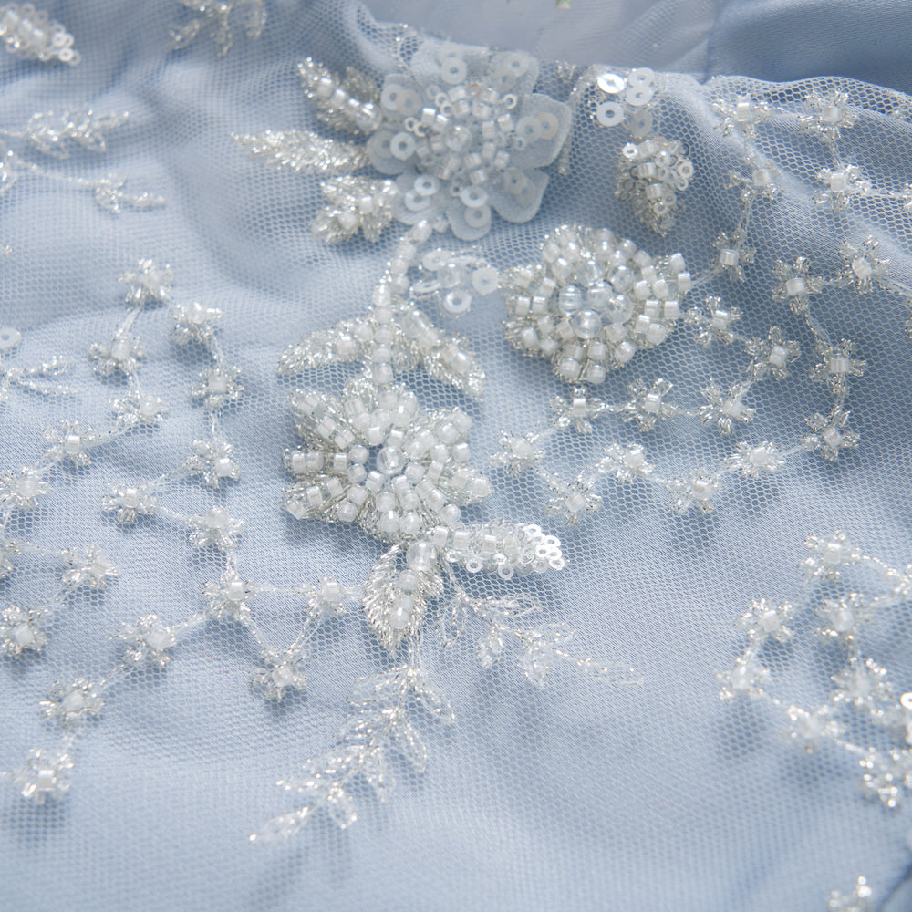 Close-up of light blue fabric with white beaded floral embroidery, sequins, and delicate lace detailing.