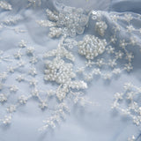 Close-up of light blue fabric with white beaded floral embroidery, sequins, and delicate lace detailing.