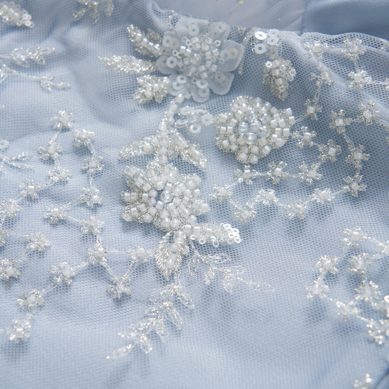 Close-up of light blue fabric with white beaded floral embroidery, sequins, and delicate lace detailing.