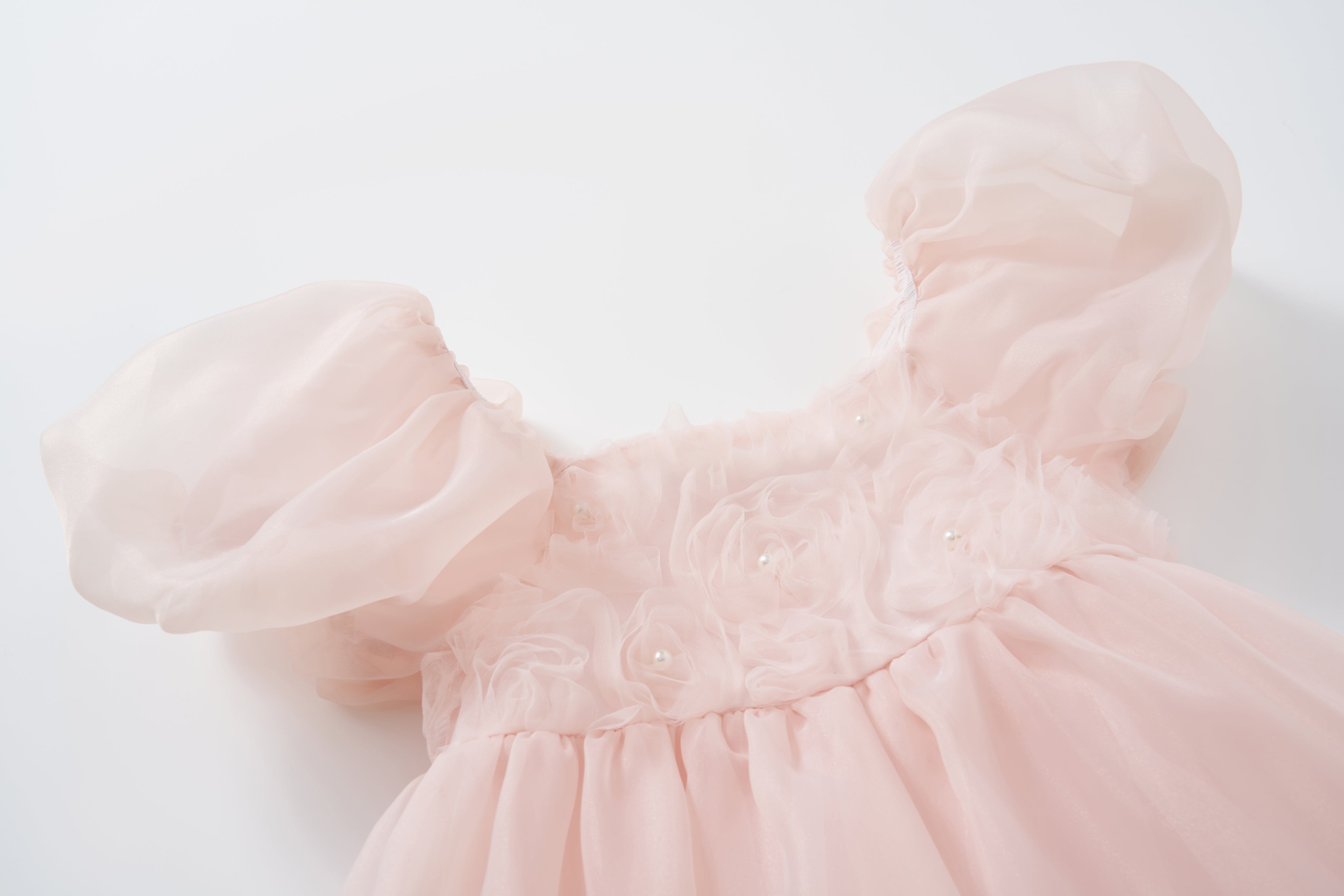 The Blush Whisper Dress by KIKI and JOJO features a princess-style design in blush pink, with sheer puff sleeves and a floral textured bodice embellished with 3D floral appliqués and tiny pearls, shown against a white background.