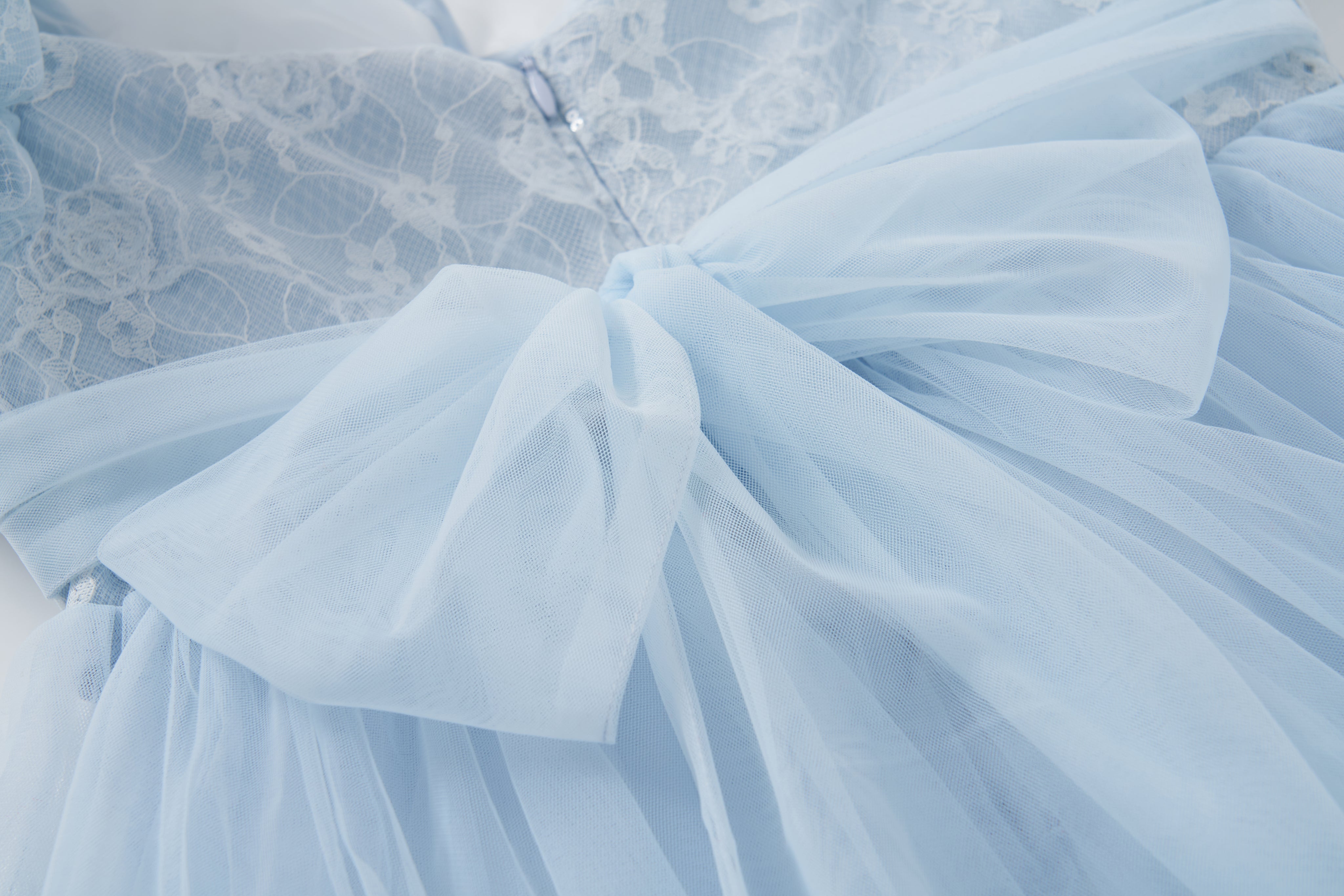 The Sky Whisper Dress by KIKI and JOJO features light blue lace and organza, a large waist bow, delicate floral lace accents, puff sleeves, and a zippered bodice.