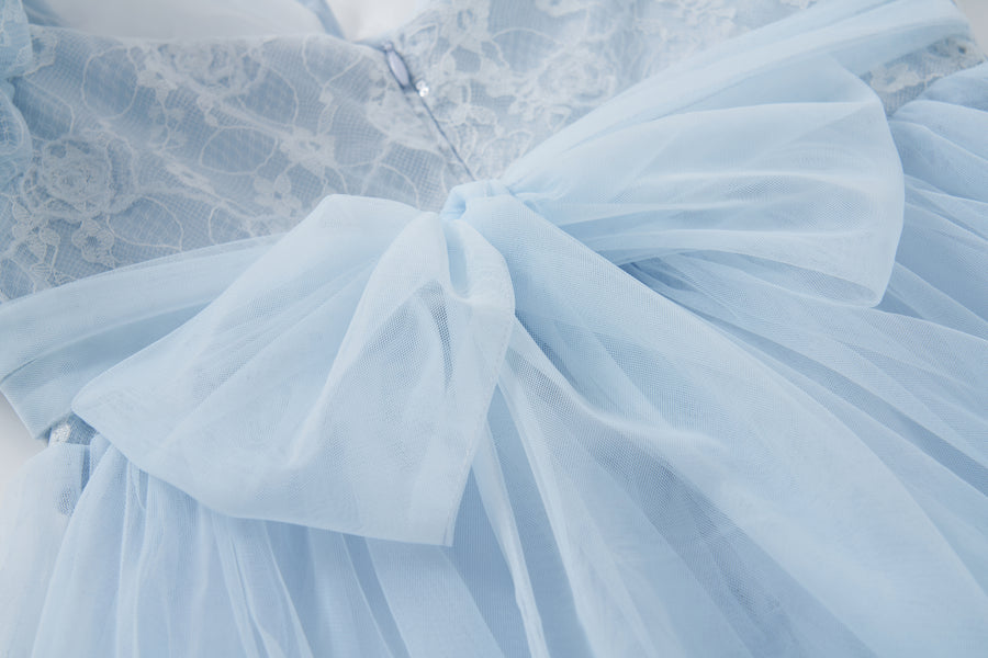 The Sky Whisper Dress by KIKI and JOJO features light blue lace and organza, a large waist bow, delicate floral lace accents, puff sleeves, and a zippered bodice.