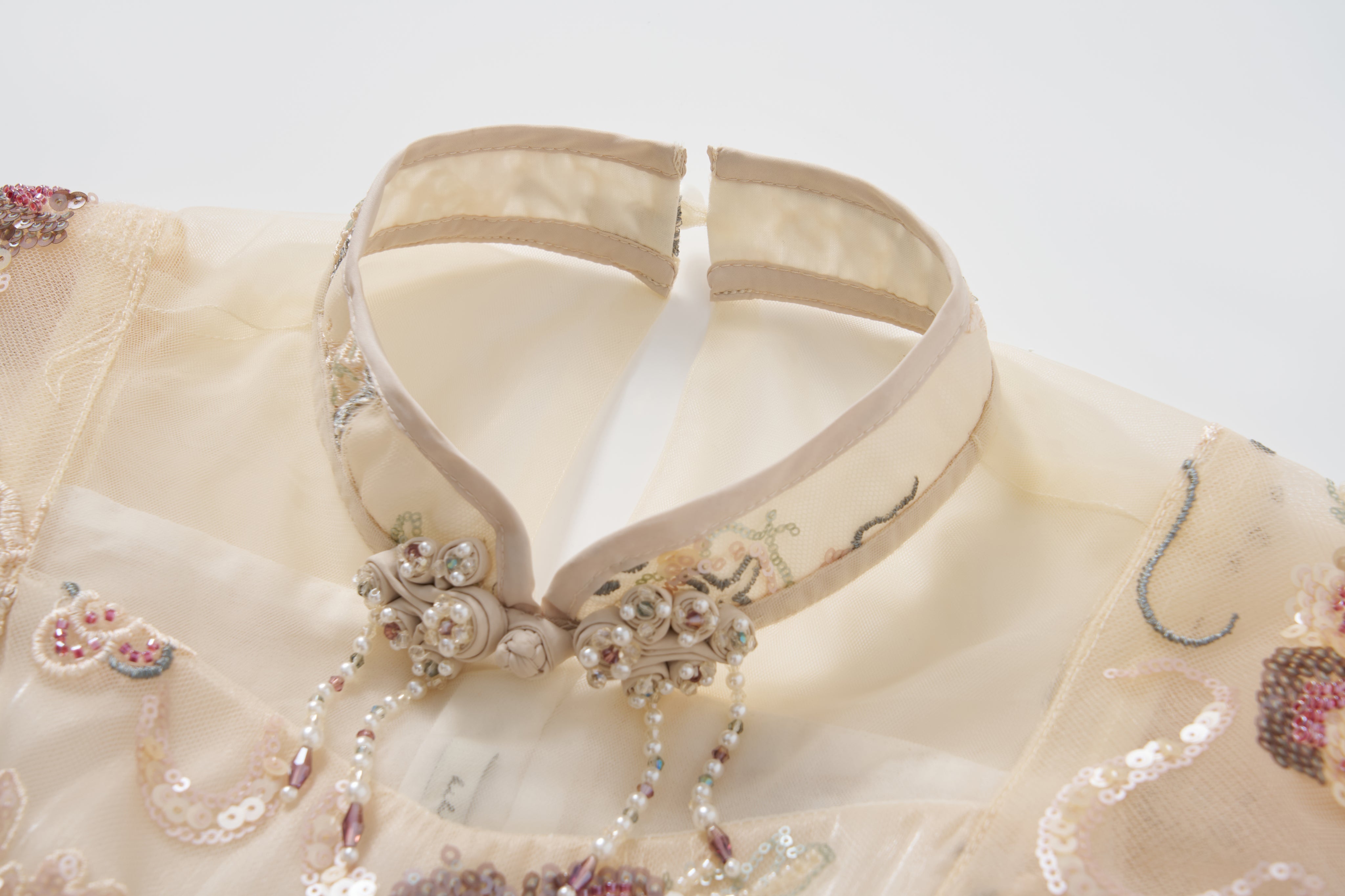 Close-up of the Jade Whisper Dress by KIKI and JOJO, a traditional cream cheongsam featuring a sheer mandarin collar, embroidered florals, sequins, and beaded neckline accents.
