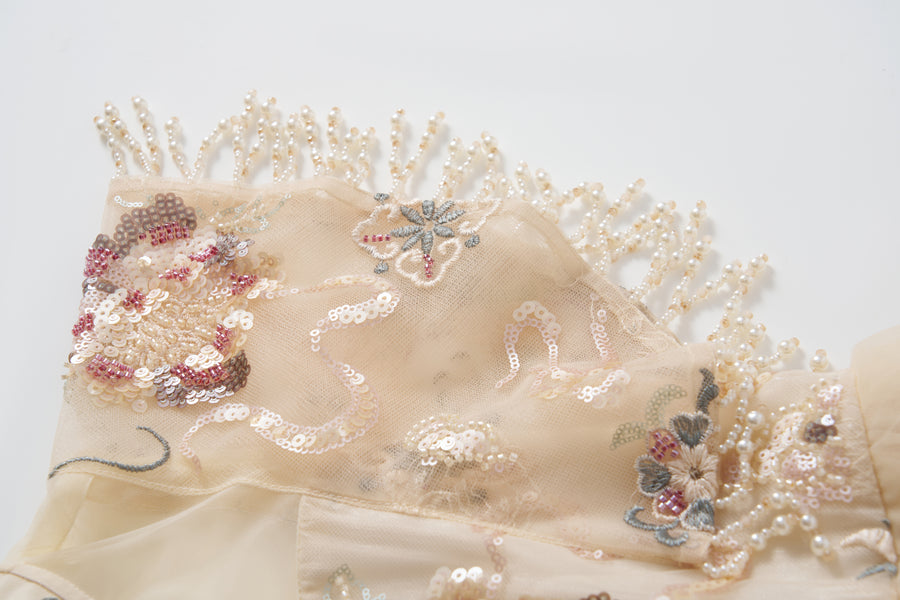 Close-up of the Jade Whisper Dress by KIKI and JOJO: beige fabric with embroidered florals, sequin details, and pearl bead fringe—perfect for an elegant cheongsam—shown on a white background.