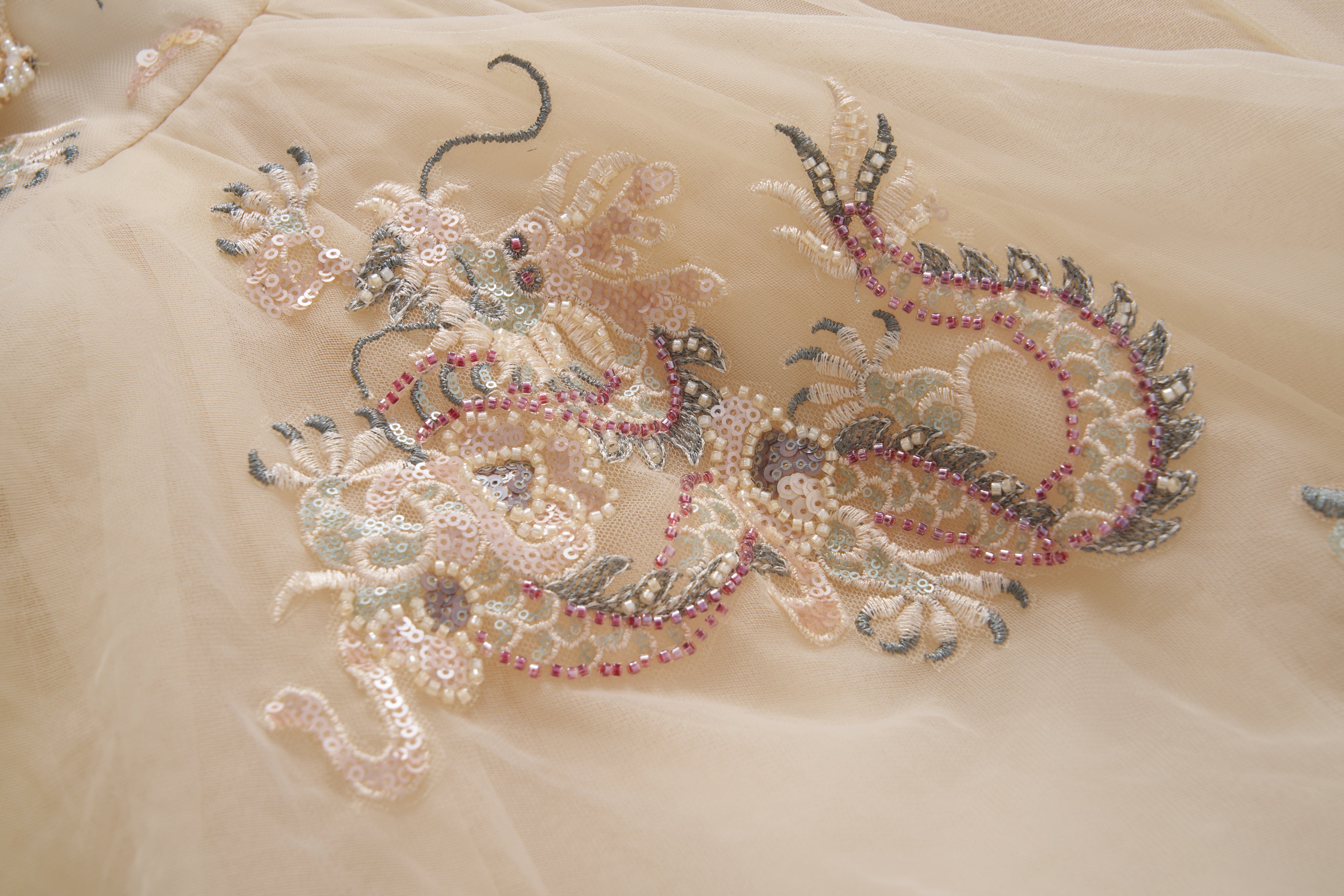 A close-up of the Jade Whisper Dress by KIKI and JOJO, showcasing its light-toned cheongsam fabric adorned with an intricate dragon motif embroidered in sequins, beads, and soft pastel hues.