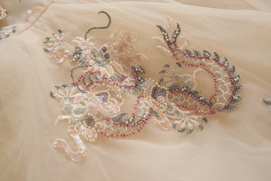 A close-up of the Jade Whisper Dress by KIKI and JOJO, showcasing its light-toned cheongsam fabric adorned with an intricate dragon motif embroidered in sequins, beads, and soft pastel hues.