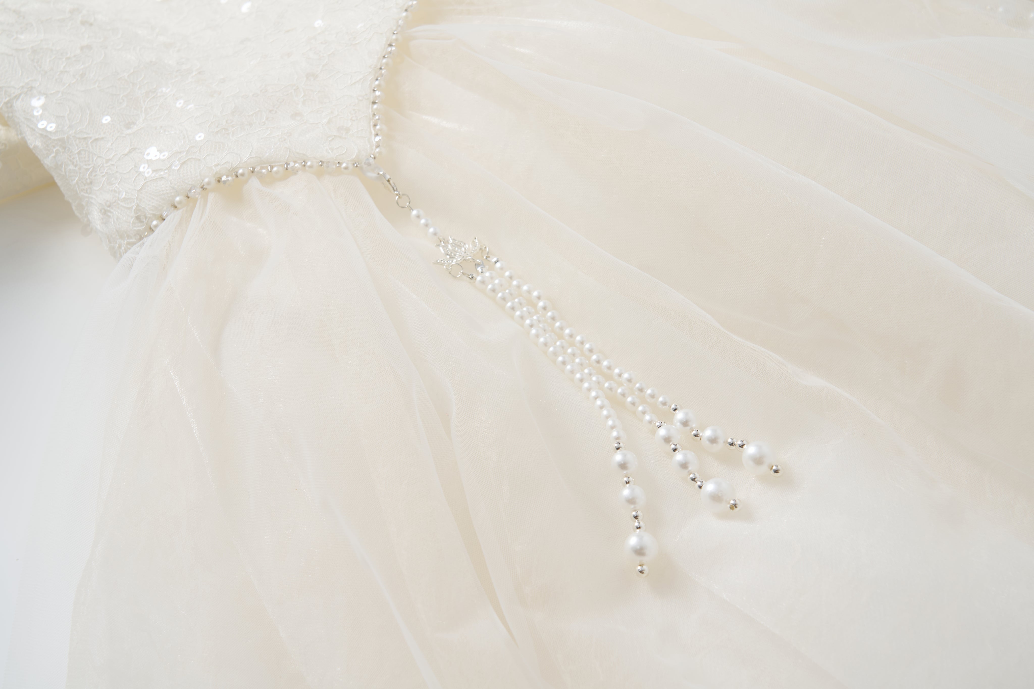 Close-up of the Orchid Whisper Dress by KIKI and JOJO, showcasing a white formal design with a sequin lace bodice and pearl bead strands cascading from the waist for a modern vintage look.