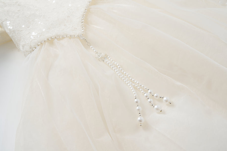Close-up of the Orchid Whisper Dress by KIKI and JOJO, showcasing a white formal design with a sequin lace bodice and pearl bead strands cascading from the waist for a modern vintage look.