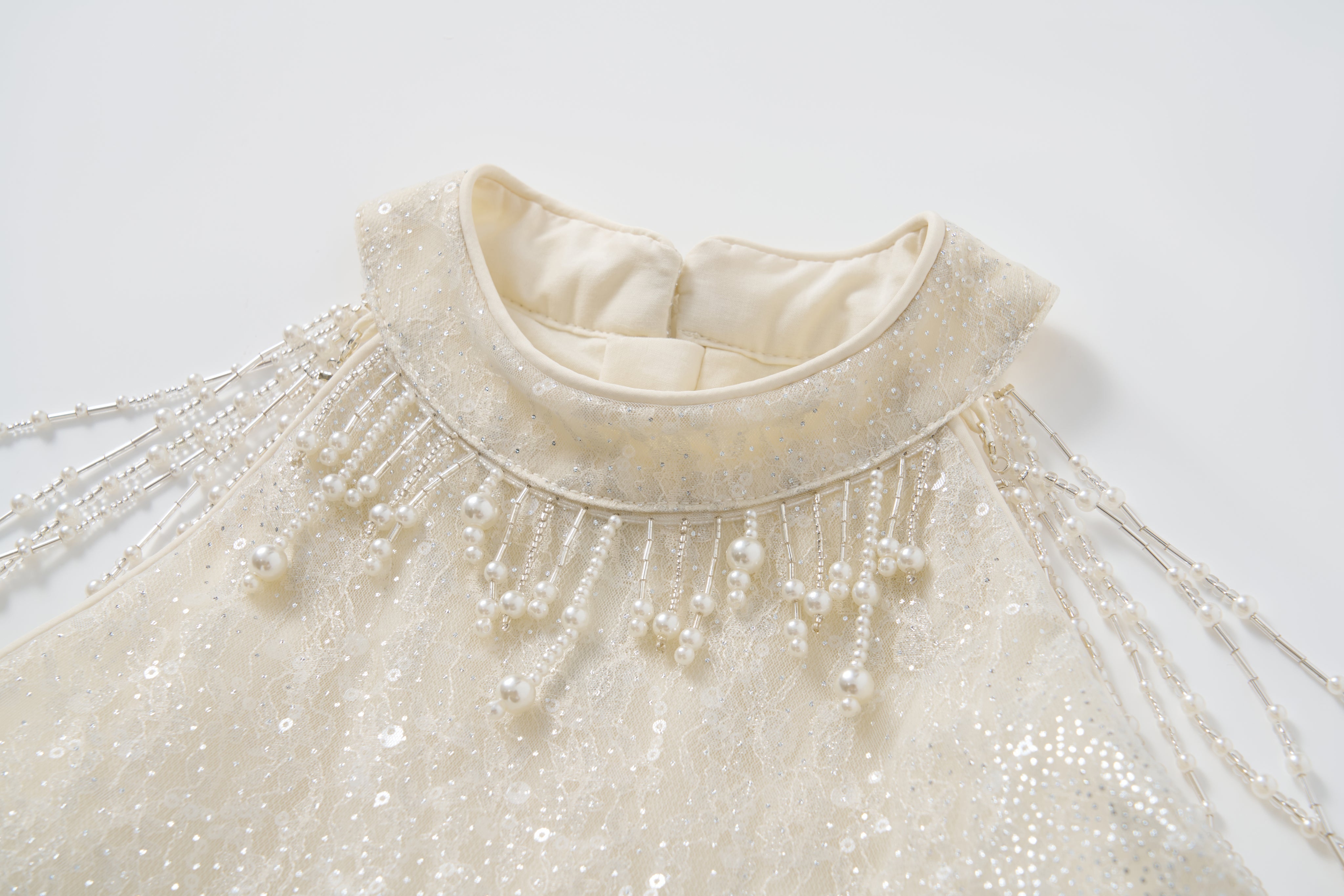 Close-up of the Gilded Whisper Dress by KIKI and JOJO—an ivory halter-neck sequin dress with a high lace neckline and beaded pearl fringe detailing on the shoulders.