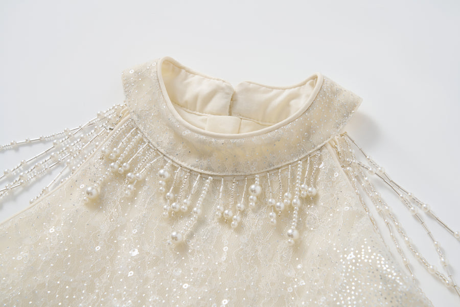 Close-up of the Gilded Whisper Dress by KIKI and JOJO—an ivory halter-neck sequin dress with a high lace neckline and beaded pearl fringe detailing on the shoulders.