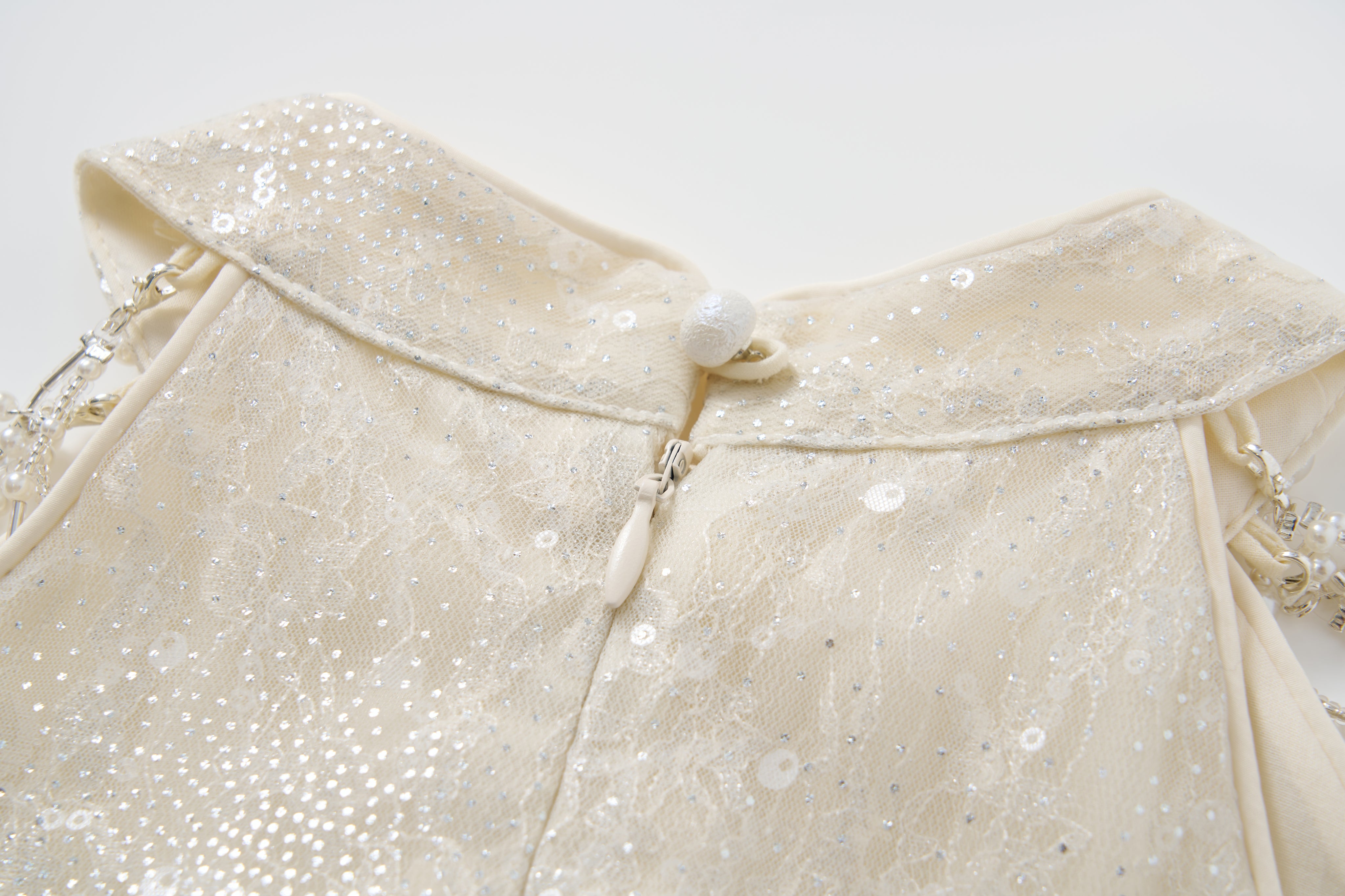 Close-up of the back of the KIKI and JOJO Gilded Whisper Dress: a cream halter-neck sequin dress with lace fabric, zipper, fabric-covered button, and beaded shoulder straps.