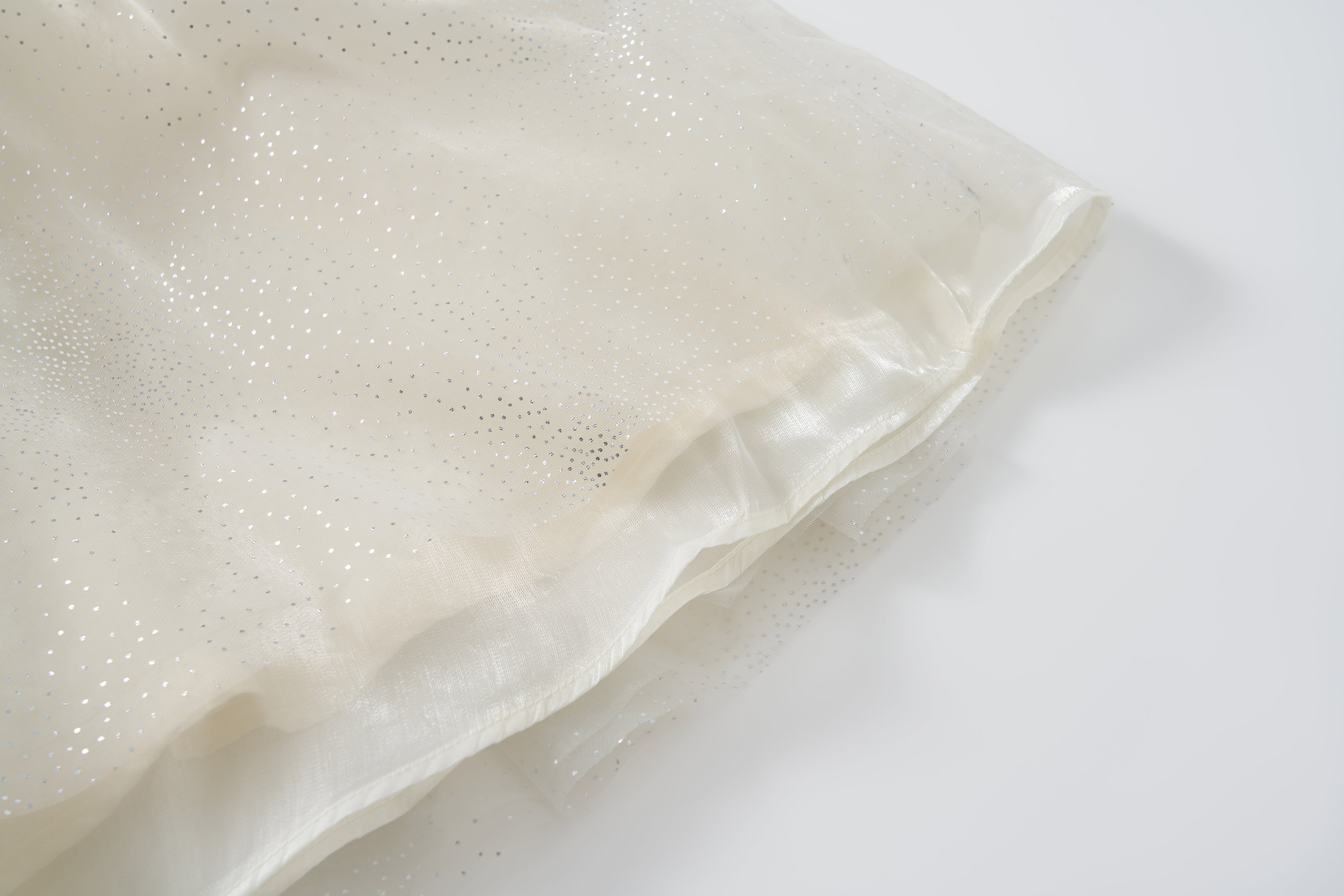 Close-up of the Gilded Whisper Dress by KIKI and JOJO, featuring sheer off-white fabric with a shimmery, dotted pattern reminiscent of beige tulle, draped elegantly on a white surface.