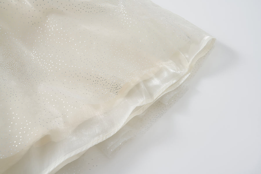 Close-up of the Gilded Whisper Dress by KIKI and JOJO, featuring sheer off-white fabric with a shimmery, dotted pattern reminiscent of beige tulle, draped elegantly on a white surface.