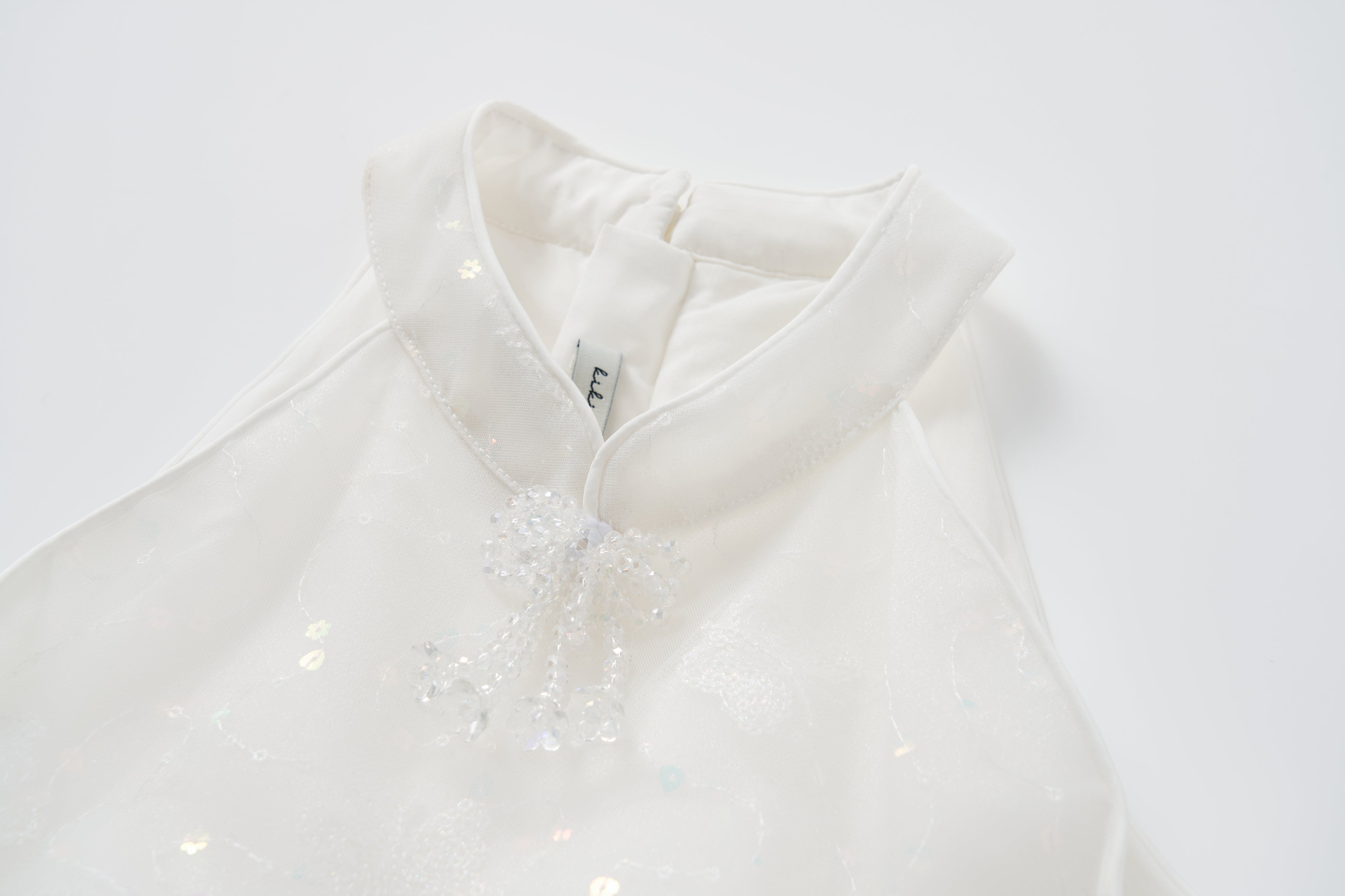 A close-up of the Butterfly Whispers Dress by KIKI and JOJO, featuring a white butterfly halter design, stand-up collar, beaded decorative knot, and a visible clothing tag.