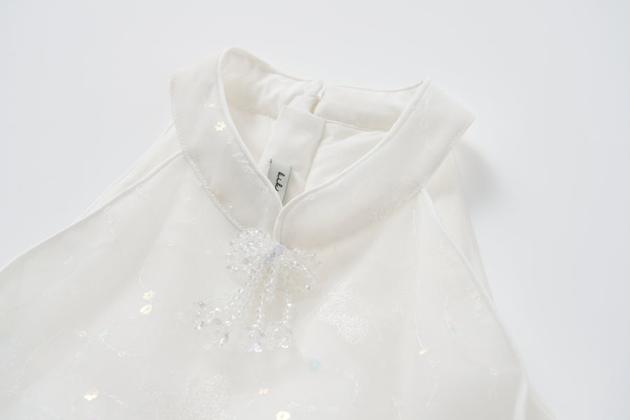 A close-up of the Butterfly Whispers Dress by KIKI and JOJO, featuring a white butterfly halter design, stand-up collar, beaded decorative knot, and a visible clothing tag.