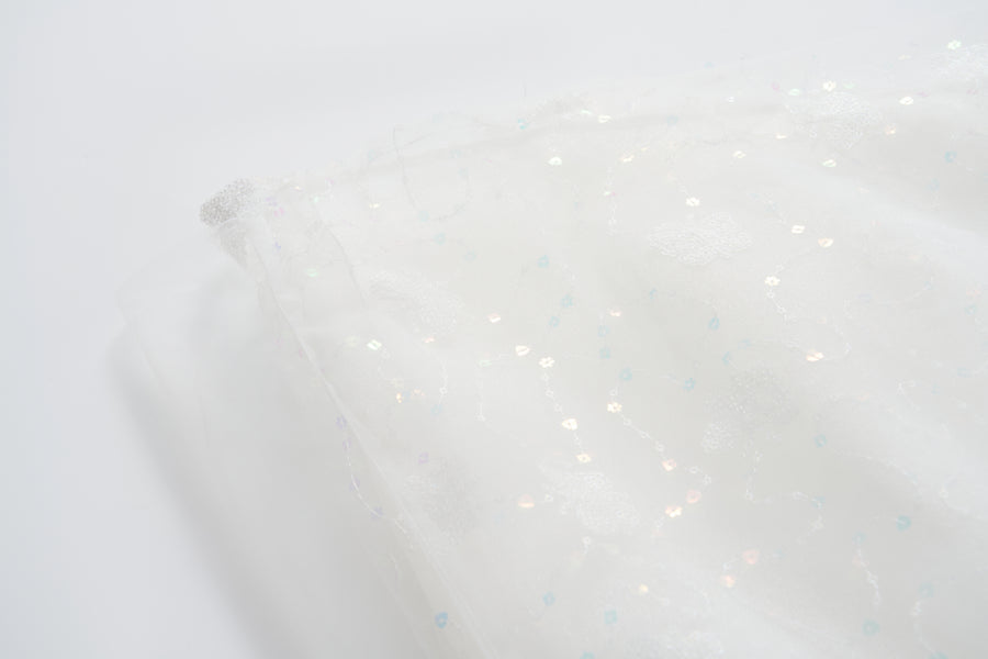 A close-up of the Butterfly Whispers Dress by KIKI and JOJO shows semi-transparent white fabric with a shiny embroidered floral pattern, capturing the delicate elegance of layered tulle, set against a plain white background.