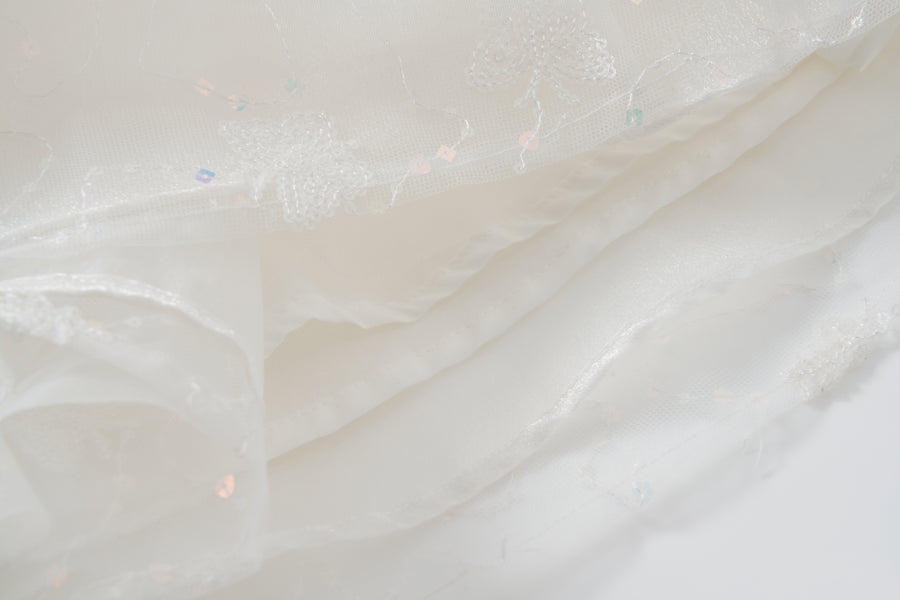 Close-up of layered white sheer fabric with embroidered floral patterns and iridescent sequins from the Butterfly Whispers Dress by KIKI and JOJO.