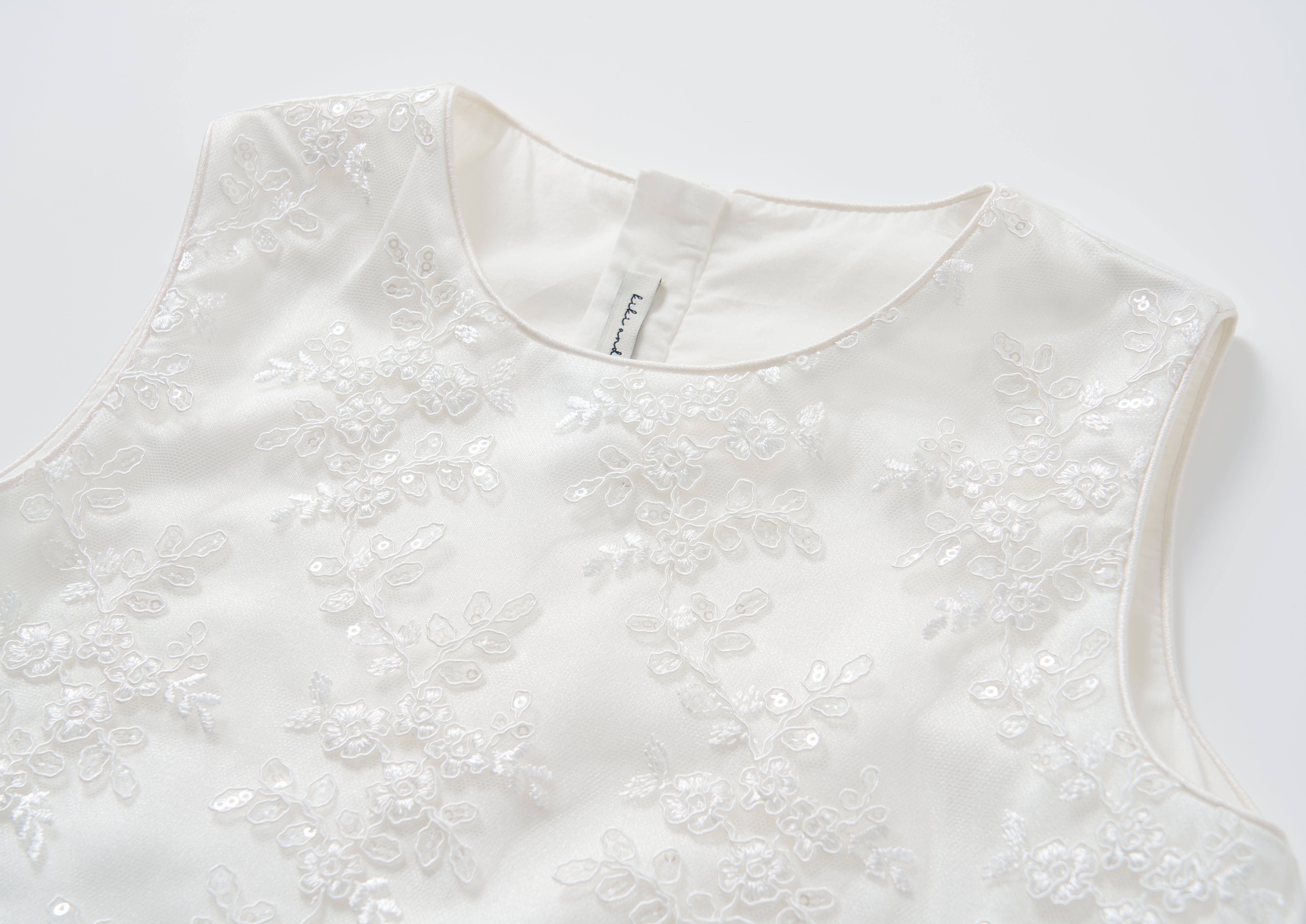 A close-up of the Twinkle Whisper Dress by KIKI and JOJO, a white A-line dress featuring floral lace embroidery and sequin details, shown on a plain background.