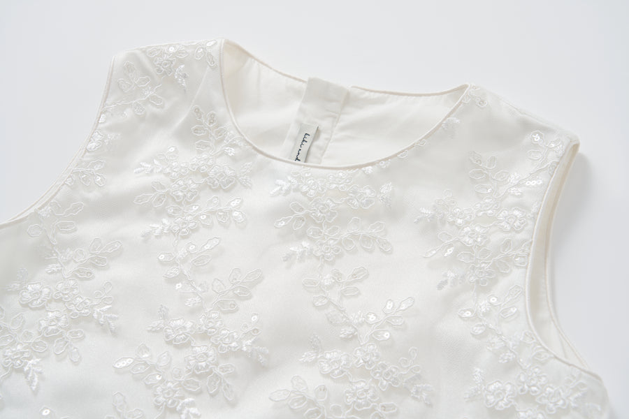 A close-up of the Twinkle Whisper Dress by KIKI and JOJO, a white A-line dress featuring floral lace embroidery and sequin details, shown on a plain background.