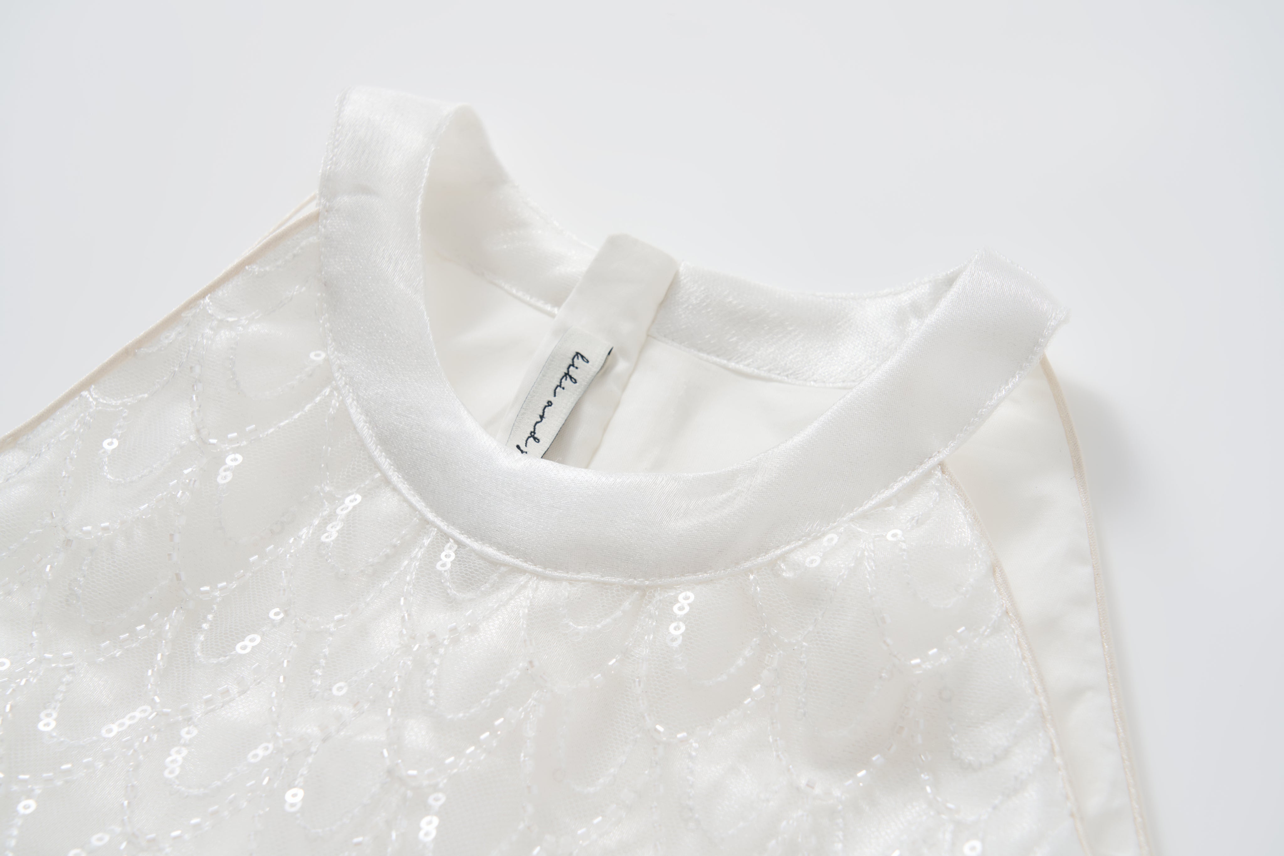 Close-up of the KIKI and JOJO Pearl Glow Whisper Dress—sleeveless with satin collar, delicate sequin patterns, and subtle beaded embroidery—laid flat on a white surface.