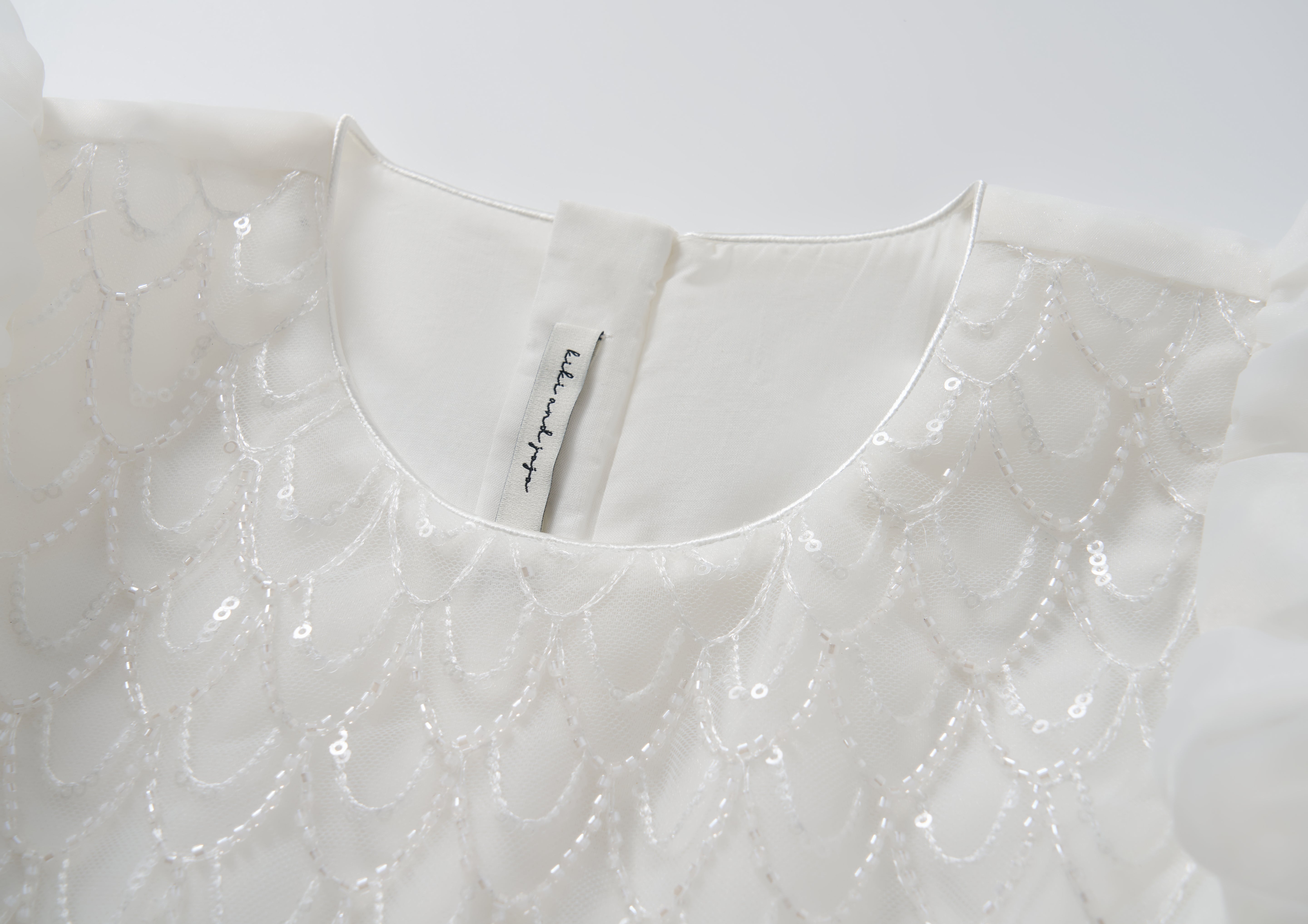 Close-up of the Ballerina Whisper Dress by KIKI and JOJO, showcasing sequin scallop patterns and beaded embroidery, with a back button closure and an inner collar fabric label.